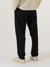 A model wears the Black Valencia Cotton Military Chino, shown from a side profile against a plain white background. The image highlights the straight-leg fit and the clean construction of the black Valencia cotton fabric. The model is wearing the trousers from the waist down, paired with a light-colored top and dark shoes.