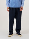A model wears the Navy Valencia Cotton Pleated Track Pant while standing against a plain white background. The image shows the trousers from the waist down, highlighting the loose, tapered fit and the single pleat at the front. The elasticated waistband and the fine, plain weave of the navy blue fabric are clearly visible. The model is also wearing a light-colored long-sleeve shirt and dark shoes.
