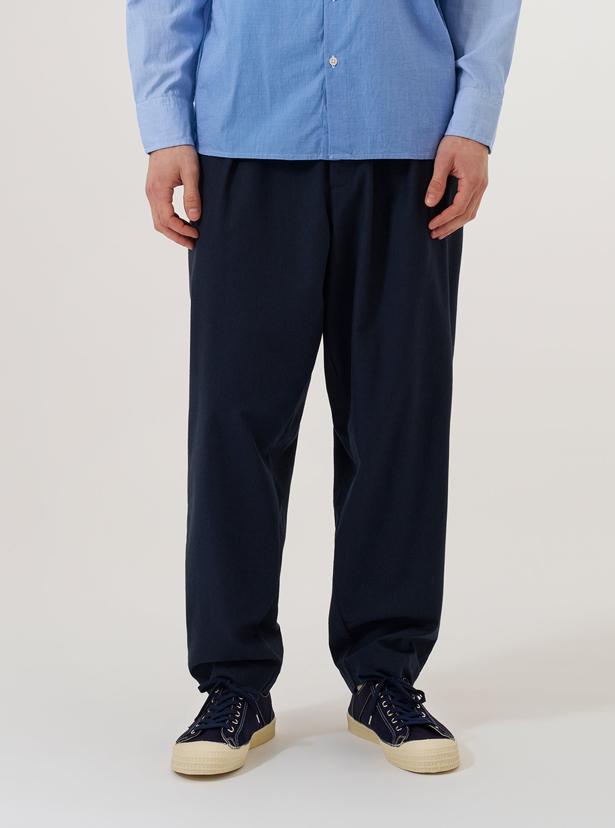 A model wears the Navy Valencia Cotton Pleated Track Pant while standing against a plain white background. The image shows the trousers from the waist down, highlighting the loose, tapered fit and the single pleat at the front. The elasticated waistband and the fine, plain weave of the navy blue fabric are clearly visible. The model is also wearing a light-colored long-sleeve shirt and dark shoes.