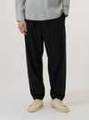 A model wears the Black Valencia Cotton Pleated Track Pant while standing against a plain white background. The image shows a front view of the trousers, highlighting the loose, tapered fit, the single pleat at the front of each leg, and the elasticated drawstring waistband. The model is wearing a white T-shirt tucked into the pants and black shoes.