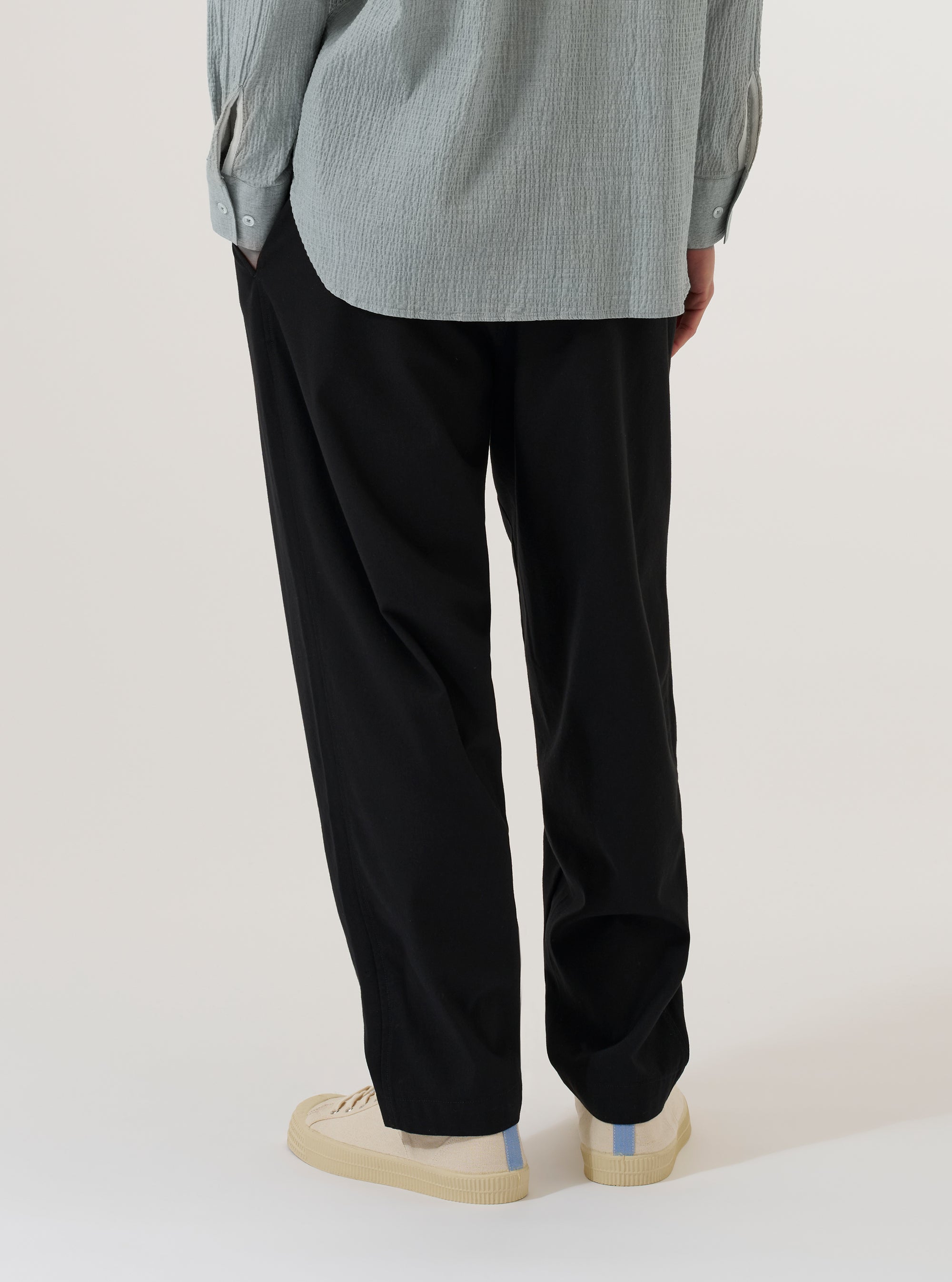A model wears the Black Valencia Cotton Pleated Track Pant, shown from the waist down against a plain white background. The image highlights the loose, tapered fit of the trousers, the single pleat at the front, and the elasticated waistband with a drawstring. The black cotton fabric has a smooth finish, and the model is wearing the pants with a white t-shirt and white socks.