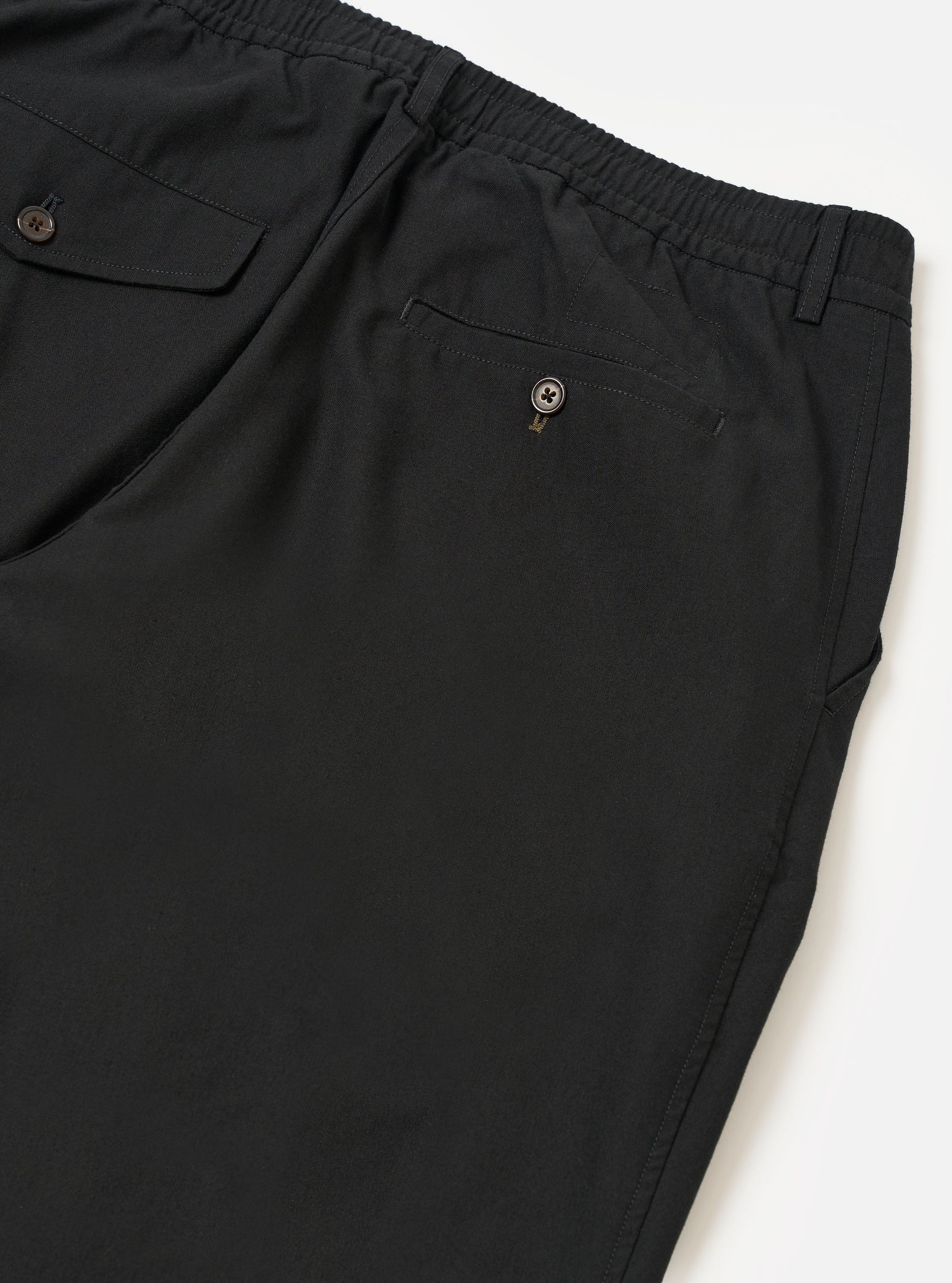 The Black Valencia Cotton Pleated Track Pant is shown from the back as a flat lay against a plain white background. The image highlights the elasticated waistband and two rear pockets: one with a buttoned flap and one buttoned jetted pocket. The black Valencia cotton fabric features a fine, plain weave.