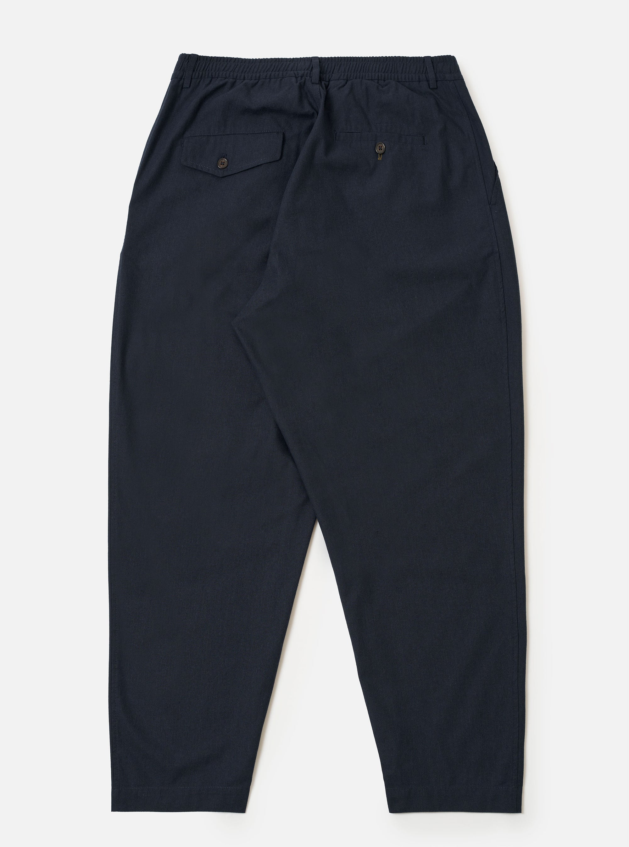 The Navy Valencia Cotton Pleated Track Pant is shown from the back as a flat lay against a plain white background. The image highlights the two rear pockets, including one with a buttoned flap and one button jetted pocket, and the elasticated waistband. The navy blue fabric has a fine, plain weave.