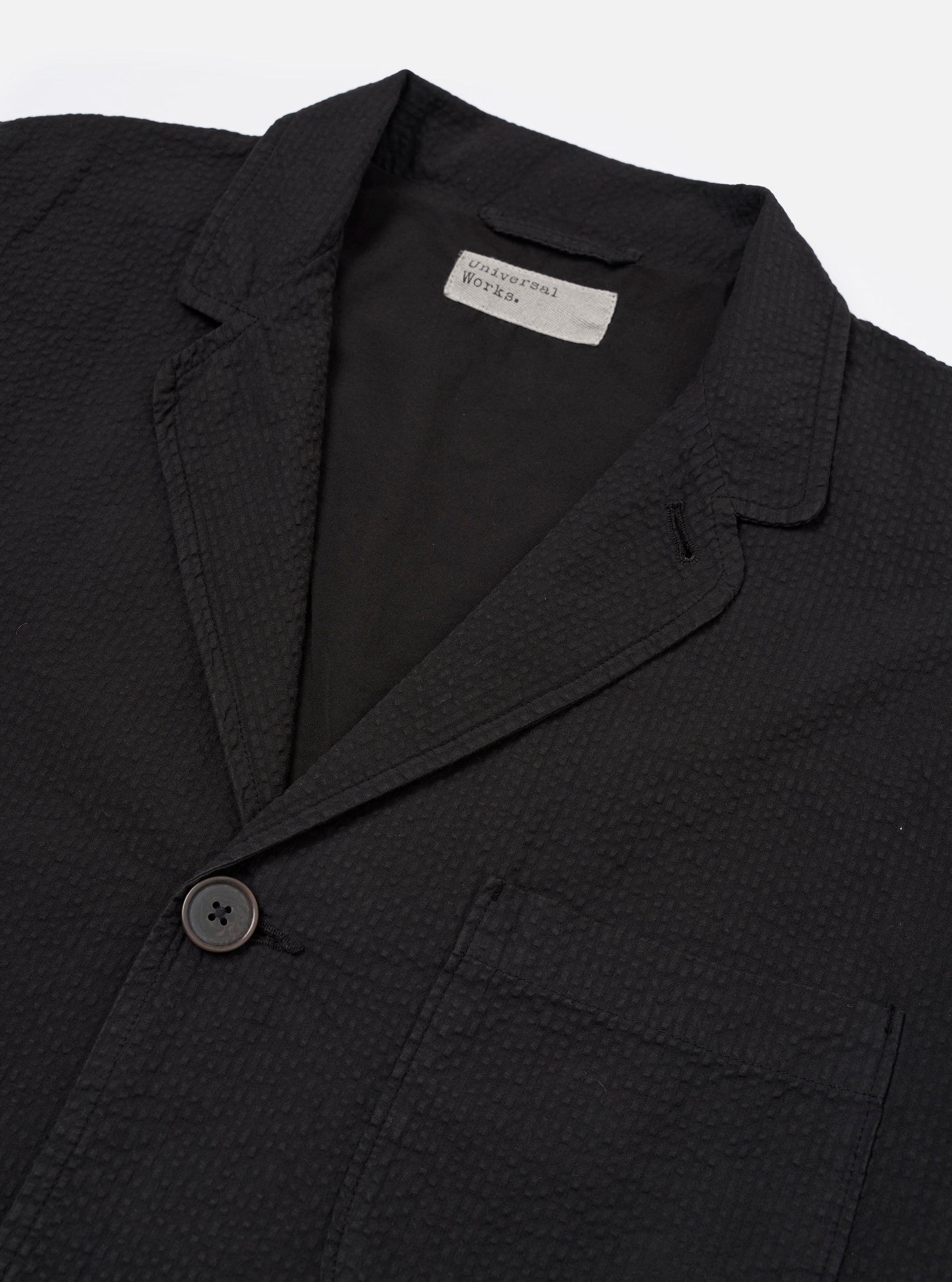 The Black Merida Seersucker Three Button Jacket is shown from the back as a flat lay against a plain white background. The image highlights the jacket's unstructured design, a central vertical seam, and the textured, puckered finish of the black seersucker fabric.