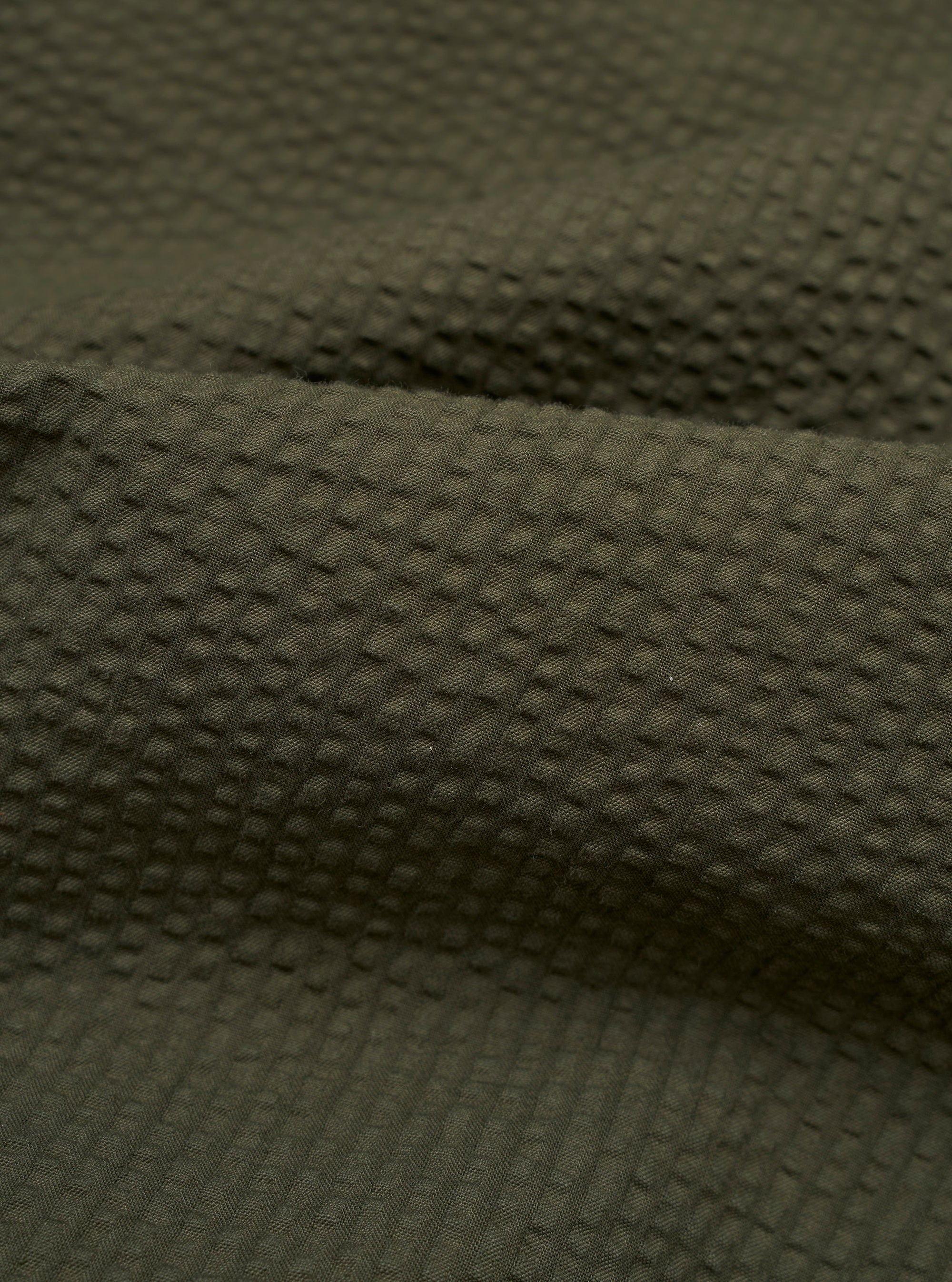 A close-up swatch image of the Olive Merida Seersucker Three Button Jacket displays the fabric's distinct puckered texture in a solid olive green color. The weave features alternating smooth and raised rows characteristic of seersucker.