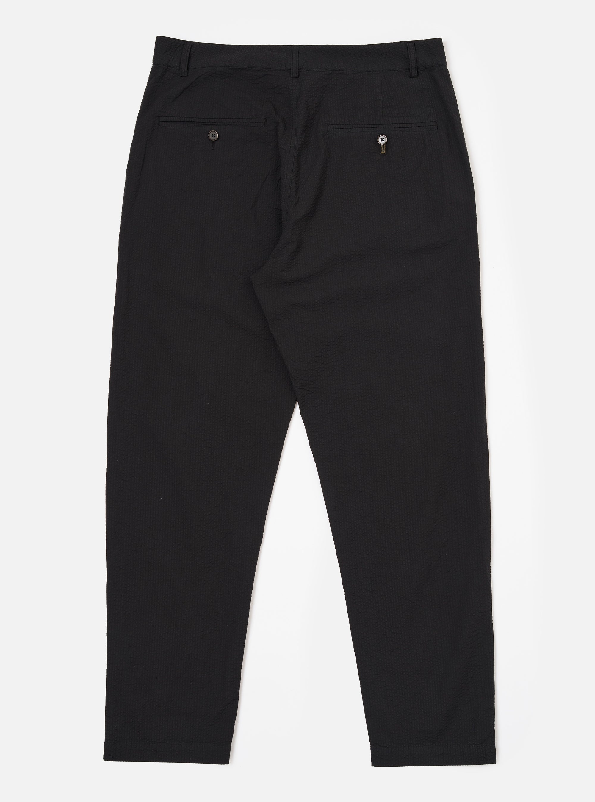 The Black Merida Seersucker Military Chino is shown from the back as a flat lay against a plain white background. The image highlights the two buttoned flap back pockets and the continuous Hollywood-style waistband. The black cotton fabric has a visible puckered seersucker texture.