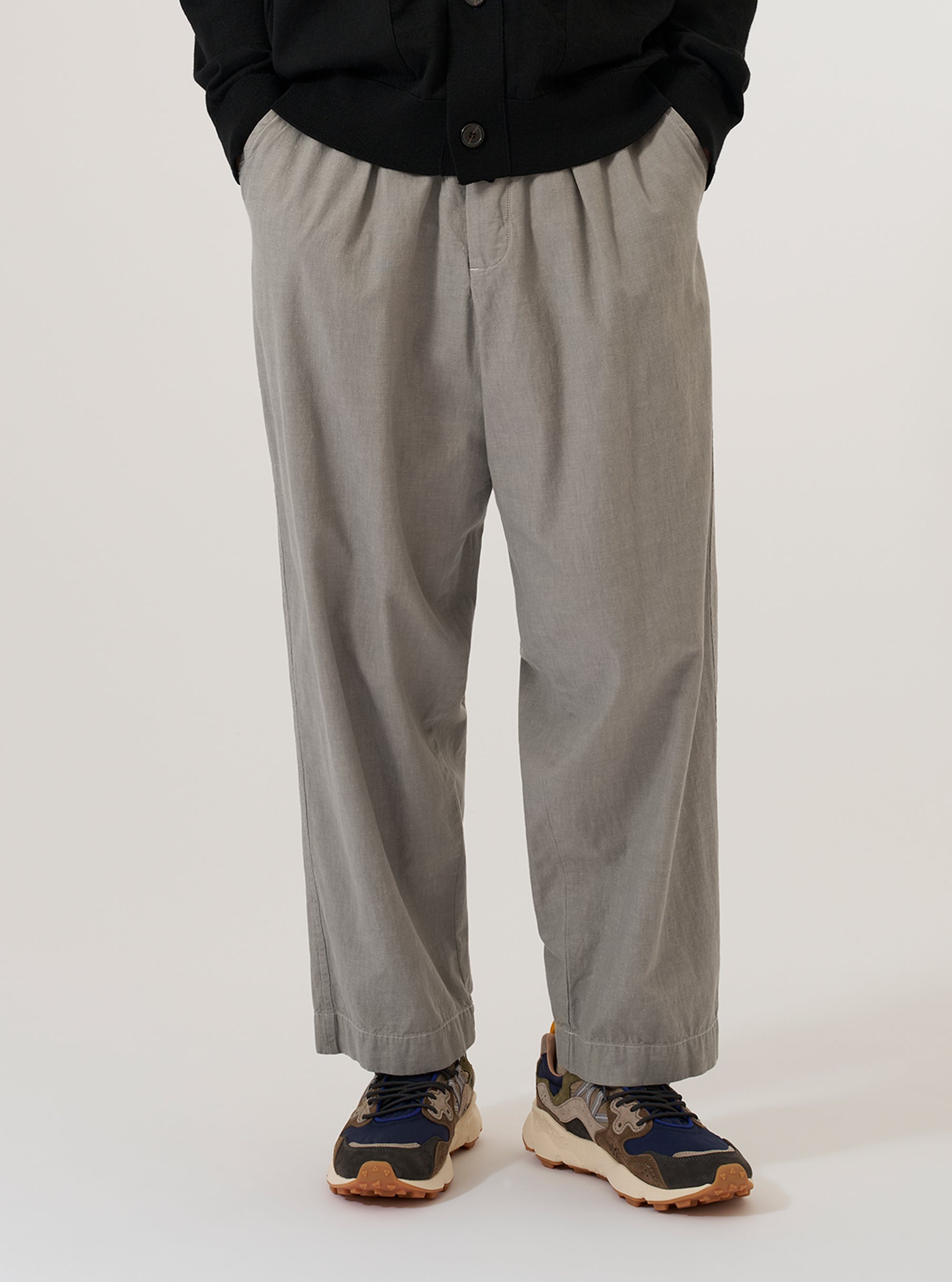 A model wears the Grey Izumi Cotton Oxford Pant, standing against a plain white background. The trousers feature a wide-leg, loose fit with distinct front pleats and an adjustable drawstring at the waist. The garment is made from a light grey cotton fabric with a visible texture.