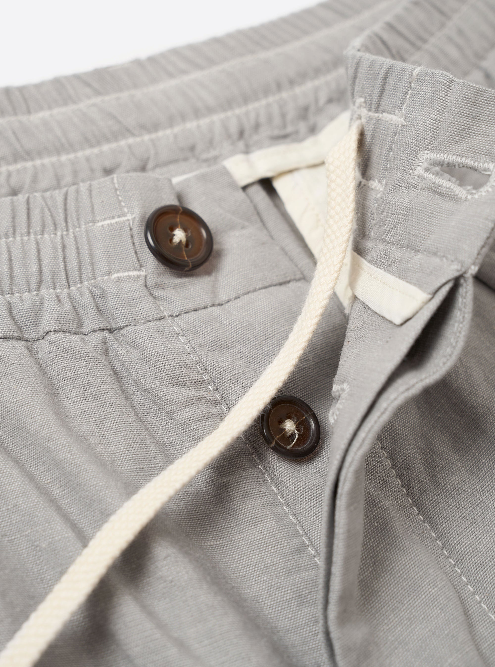 The Grey Izumi Cotton Oxford Pant is shown from the back as a flat lay against a plain white background. The image highlights the elasticated waistband and the two distinct rear pockets: one with a buttoned flap and one button jetted pocket. The trousers are a solid grey color and feature a wide leg cut.
