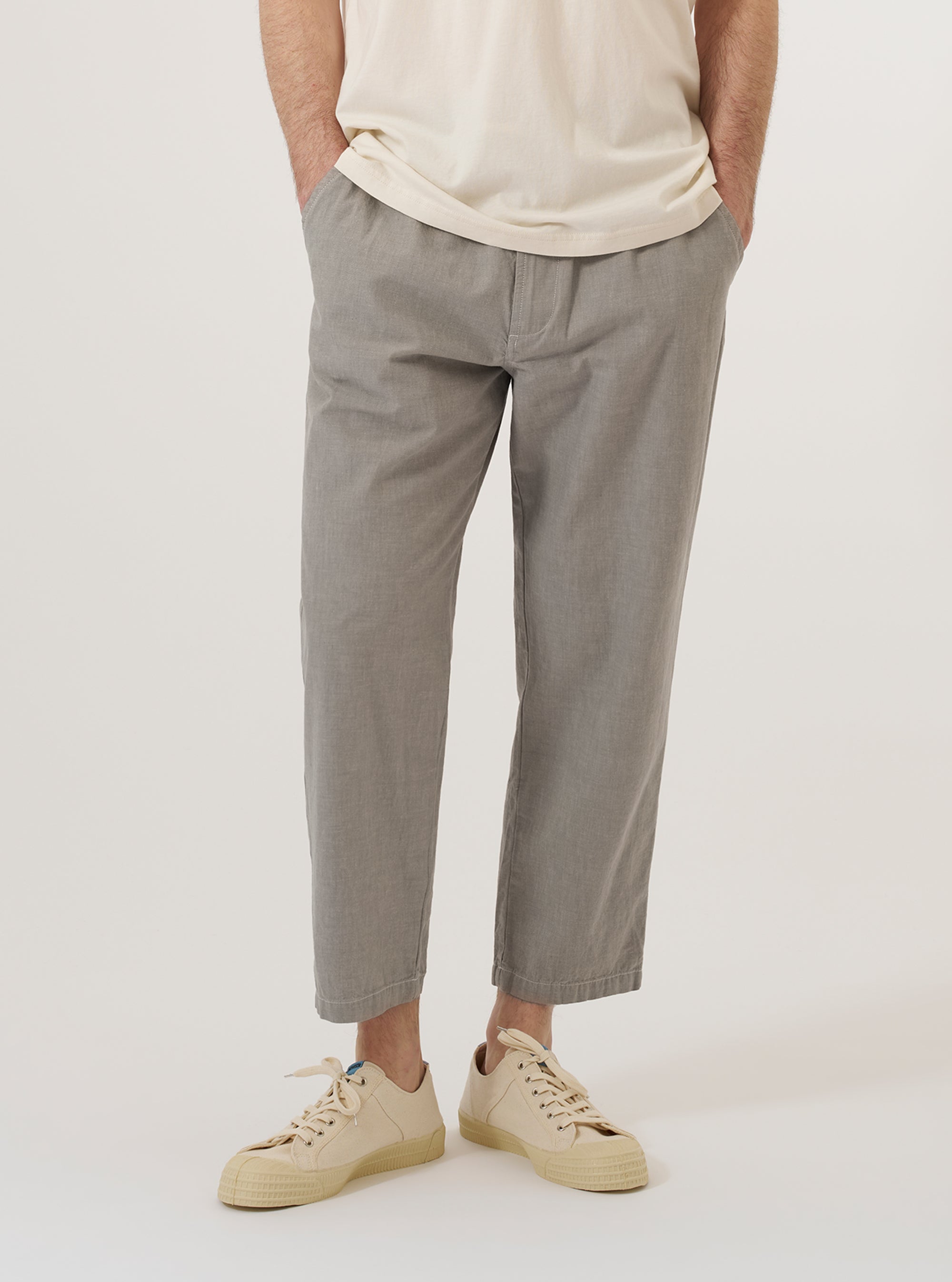 A model wears the Grey Izumi Cotton Hi Water Trouser against a plain, neutral background. The trousers feature a slim, cropped fit that ends above the ankle and an elasticated waistband with an adjustable drawstring. The grey cotton fabric has a smooth texture. The model is shown from the waist down, highlighting the garment's silhouette and length.