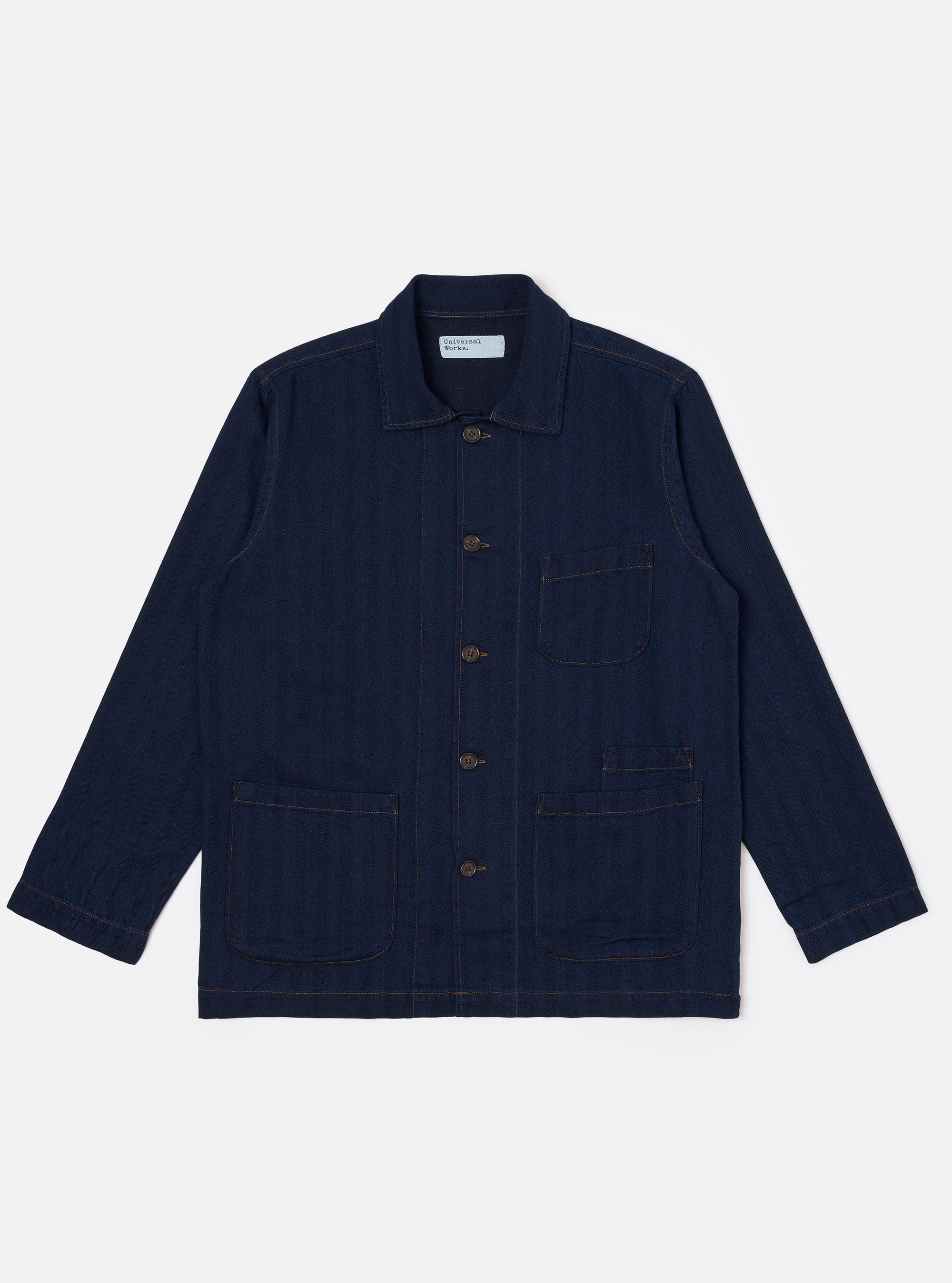 The Indigo Recycled Herringbone Bakers Overshirt is shown as a flat lay against a plain white background. The overshirt features a one-piece collar, a central button-down front, and four front patch pockets: one asymmetric pocket at the chest and three lower pockets including a small coin pocket. The indigo-dyed fabric displays a visible herringbone weave texture throughout.