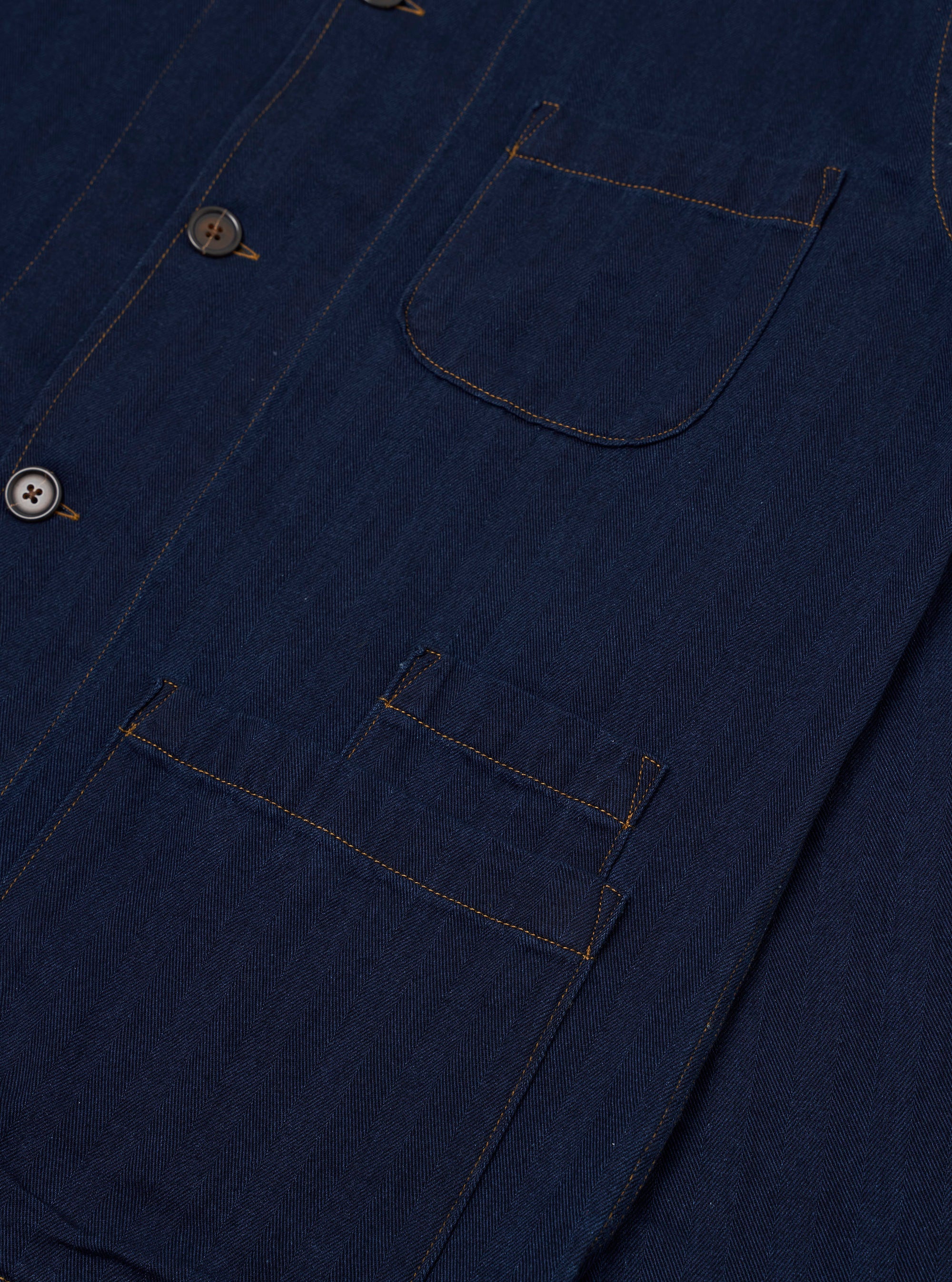 The Indigo Recycled Herringbone Bakers Overshirt is shown from the back as a flat lay against a plain white background. The image highlights the garment's textured indigo blue herringbone weave, a simple one-piece back panel, and a straight hem.