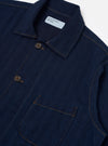 The Indigo Recycled Herringbone Bakers Overshirt is shown from the back as a flat lay against a plain white background. The image highlights the straight hem, the simple one-piece back panel, and the textured indigo blue herringbone weave of the recycled cotton fabric.