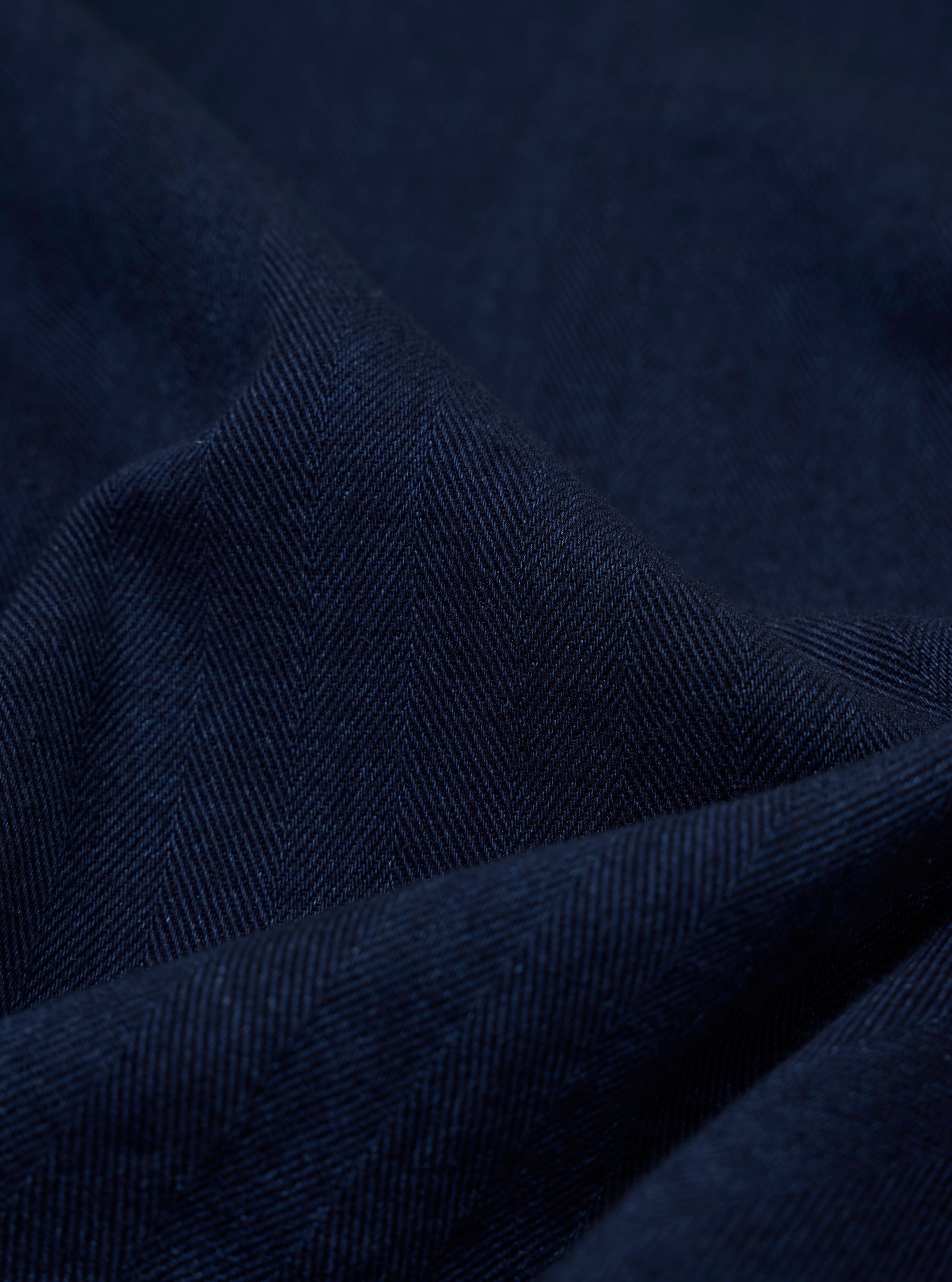 A close-up swatch image of the Indigo Recycled Herringbone Bakers Overshirt fabric. The image focuses on the textile detail, showing a deep indigo blue color with a distinct herringbone weave pattern.