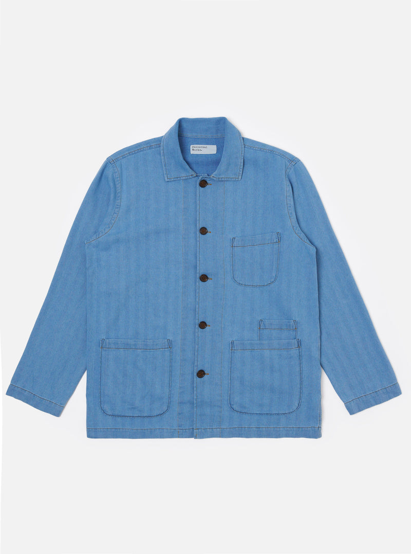 The Washed Indigo Recycled Herringbone Bakers Overshirt is shown as a flat lay against a plain white background. The overshirt features a one-piece collar, a central button-down front, and four front patch pockets: one asymmetric chest pocket, two large lower pockets, and a small coin pocket. The fabric is a washed indigo blue with a visible herringbone weave texture and a straight hem.