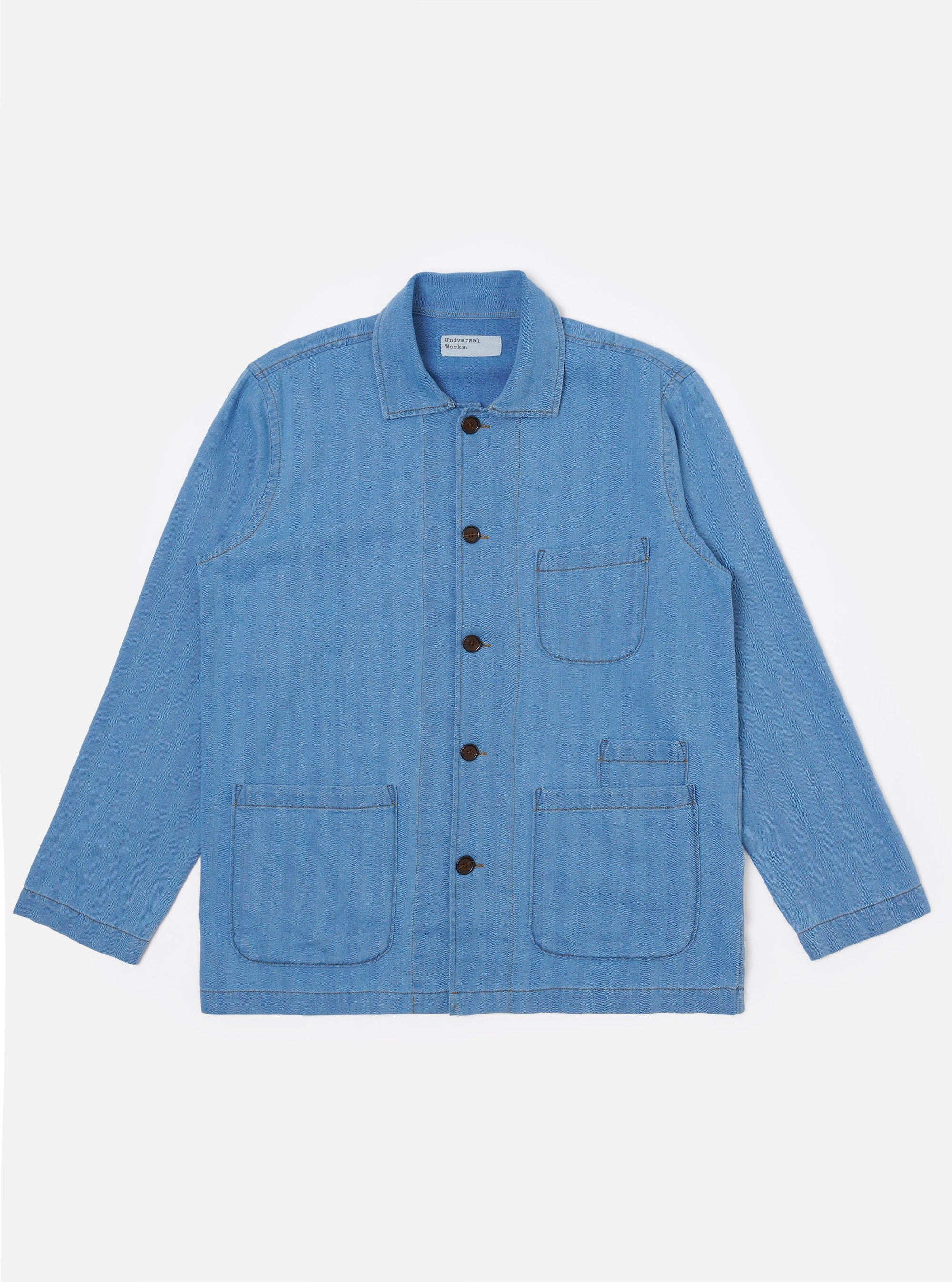 The Washed Indigo Recycled Herringbone Bakers Overshirt is shown as a flat lay against a plain white background. The overshirt features a one-piece collar, a central button-down front, and four front patch pockets: one asymmetric chest pocket, two large lower pockets, and a small coin pocket. The fabric is a washed indigo blue with a visible herringbone weave texture and a straight hem.
