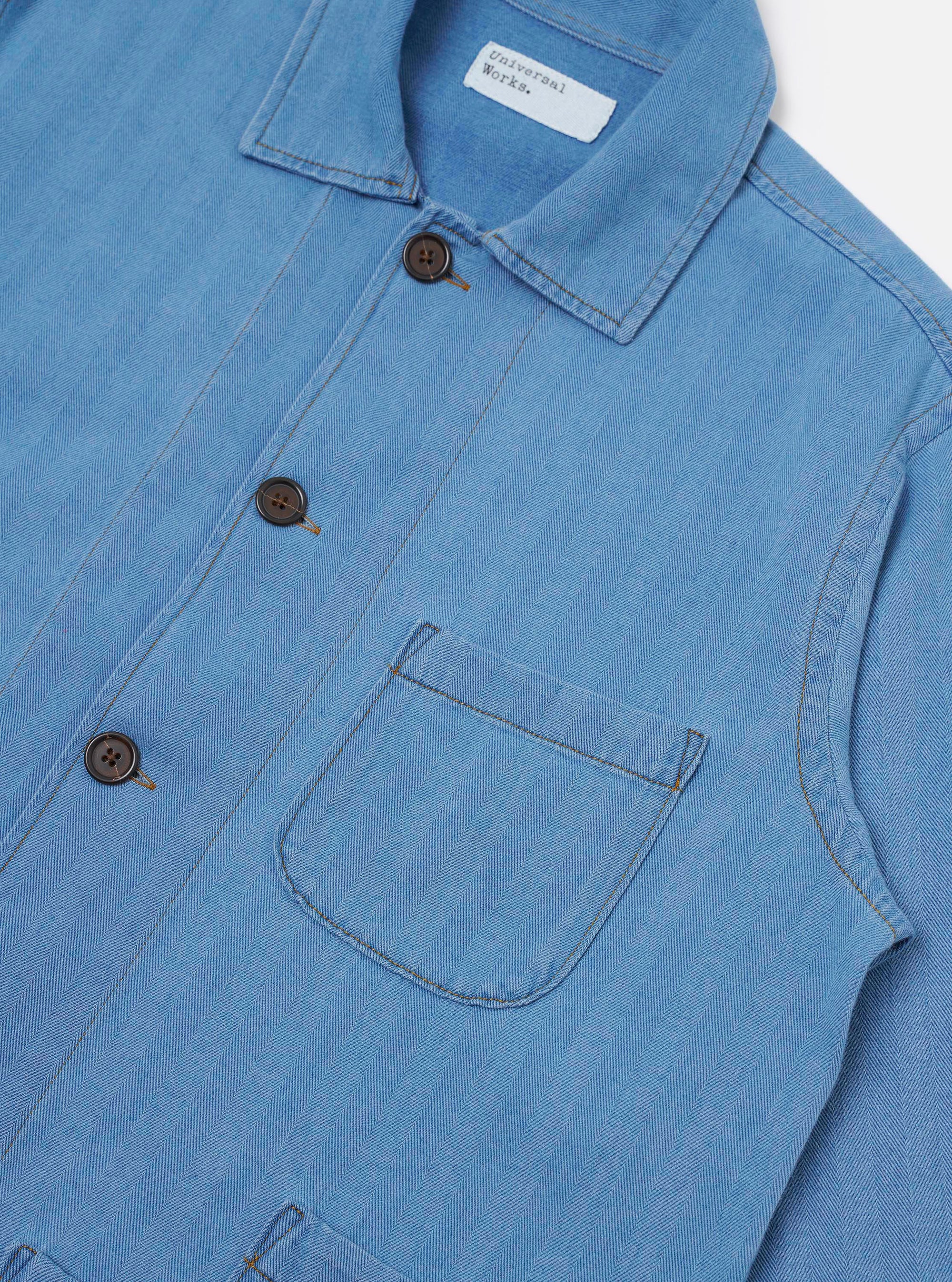 The Washed Indigo Recycled Herringbone Bakers Overshirt is shown from the back as a flat lay against a plain white background. The image highlights the clean, straight-cut back panel and the distinct V-shaped pattern of the recycled herringbone weave in a faded indigo blue.