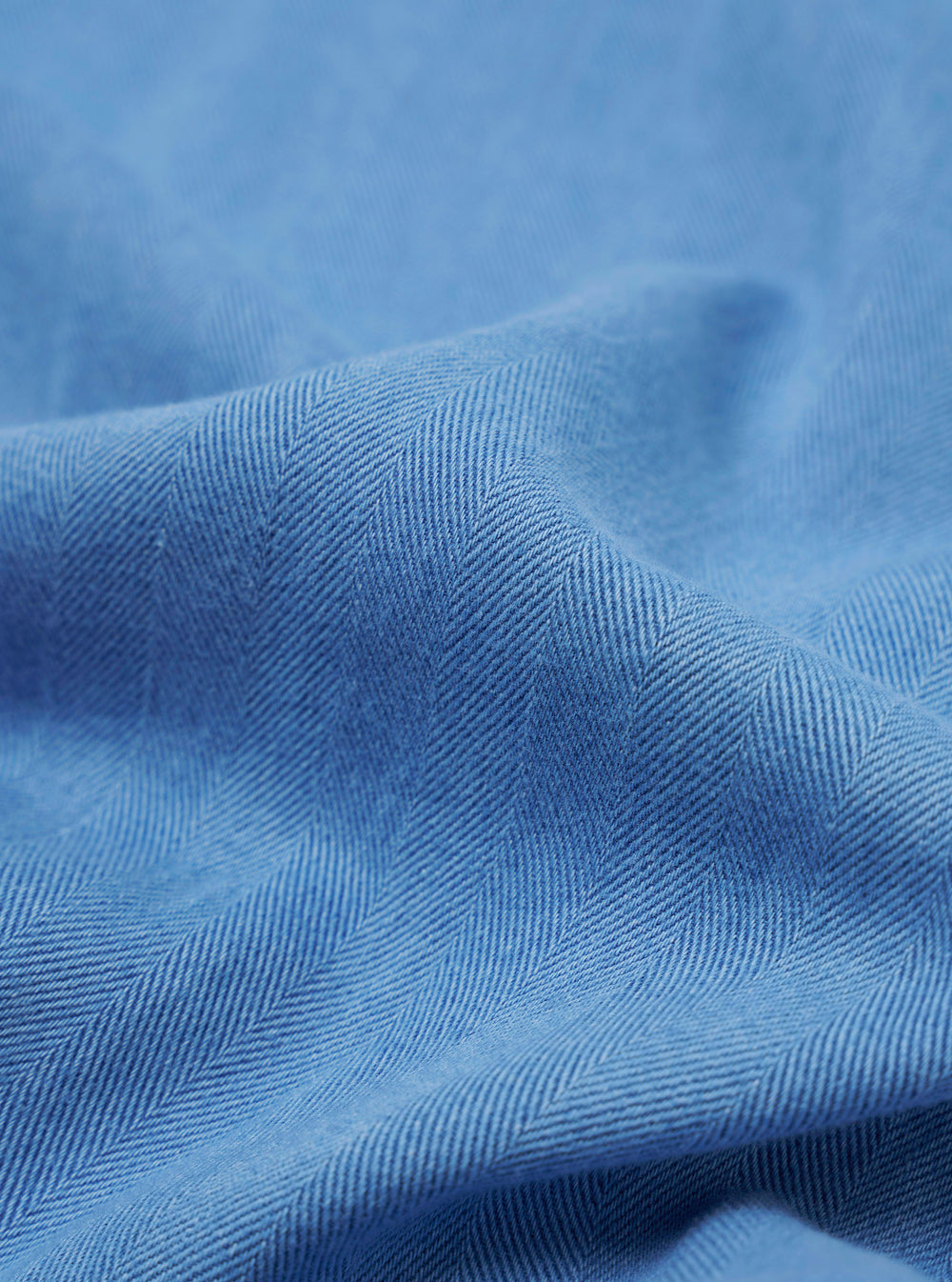 The Washed Indigo Recycled Herringbone Bakers Overshirt is shown from the back as a flat lay against a plain white background. The image highlights the clean construction of the garment's rear panel, including a straight hem and the subtle texture of the washed indigo herringbone fabric.