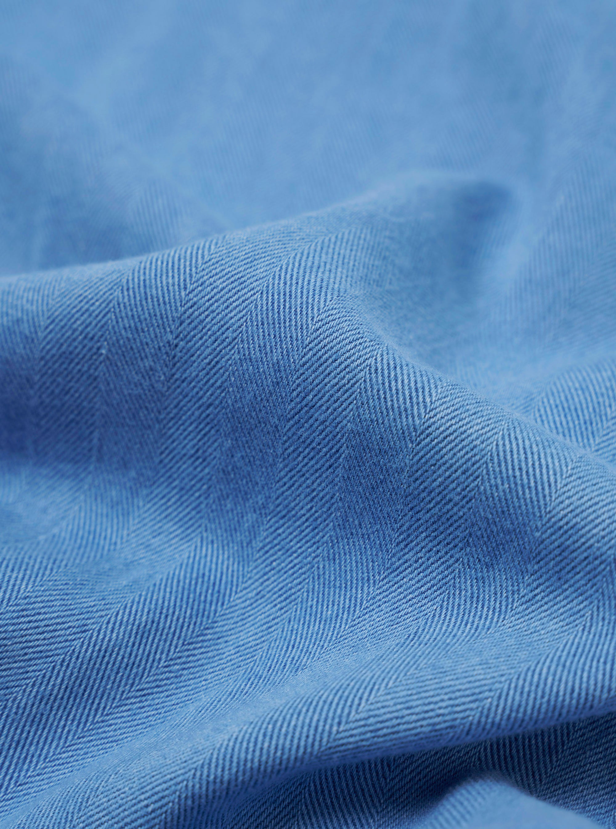 The Washed Indigo Recycled Herringbone Bakers Overshirt is shown from the back as a flat lay against a plain white background. The image highlights the clean construction of the garment's rear panel, including a straight hem and the subtle texture of the washed indigo herringbone fabric.