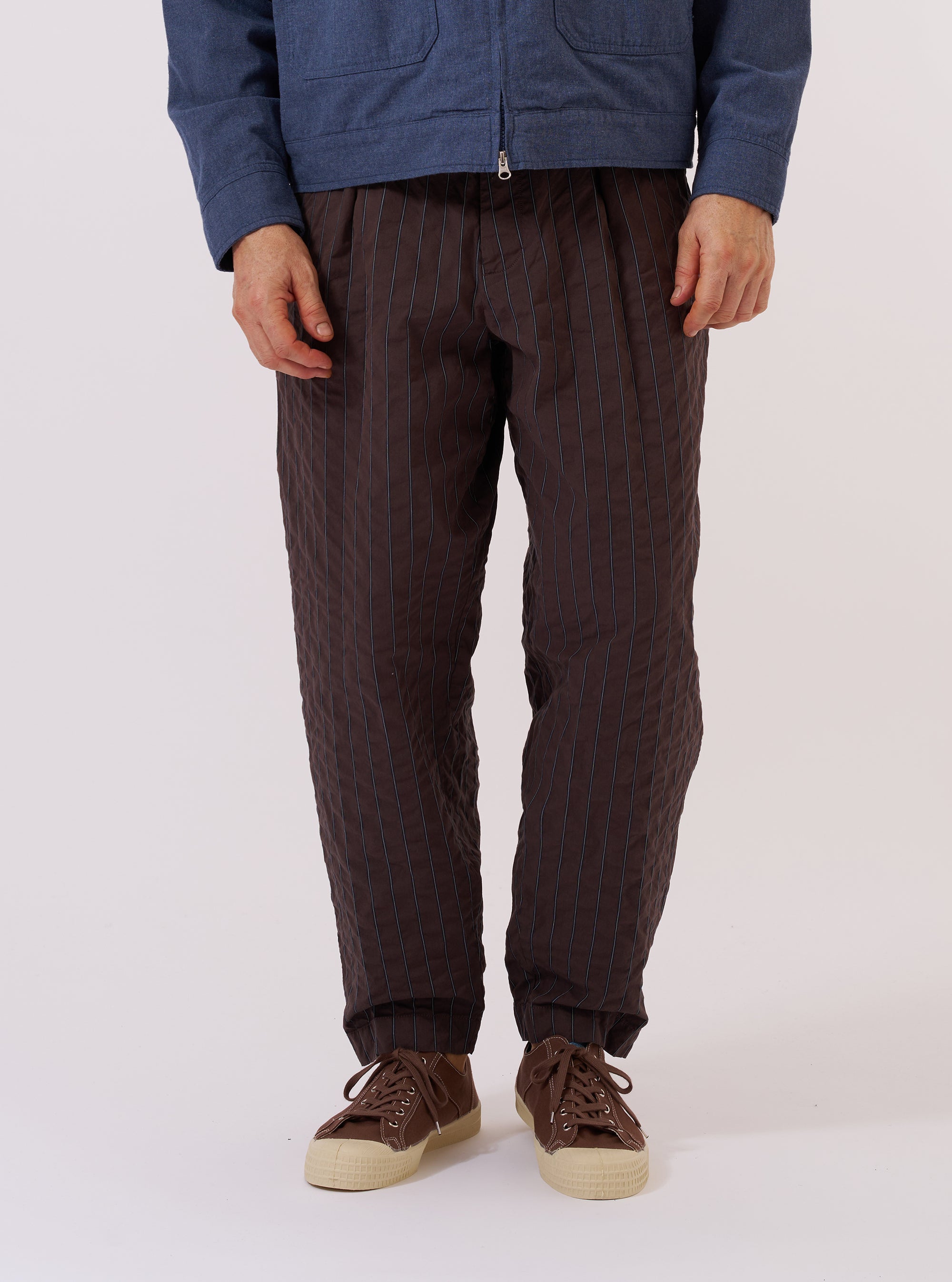 A model wears the Brown Chester Stripe Pleated Track Pant while standing against a plain white background. The image shows a front view of the trousers, highlighting the loose, tapered fit and the single pleat detail. The brown cotton seersucker fabric features a fine pinstripe pattern. The trousers are styled with the waistband visible, showcasing the elasticated construction.