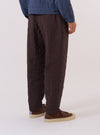 A model wears the Brown Chester Stripe Pleated Track Pant, shown from the waist down from a rear angle against a plain white background. The trousers feature a vertical brown and tan stripe pattern, a relaxed tapered fit, and a single rear patch pocket. The elasticated waistband and the textured cotton fabric are clearly visible as the model stands with one leg slightly forward.