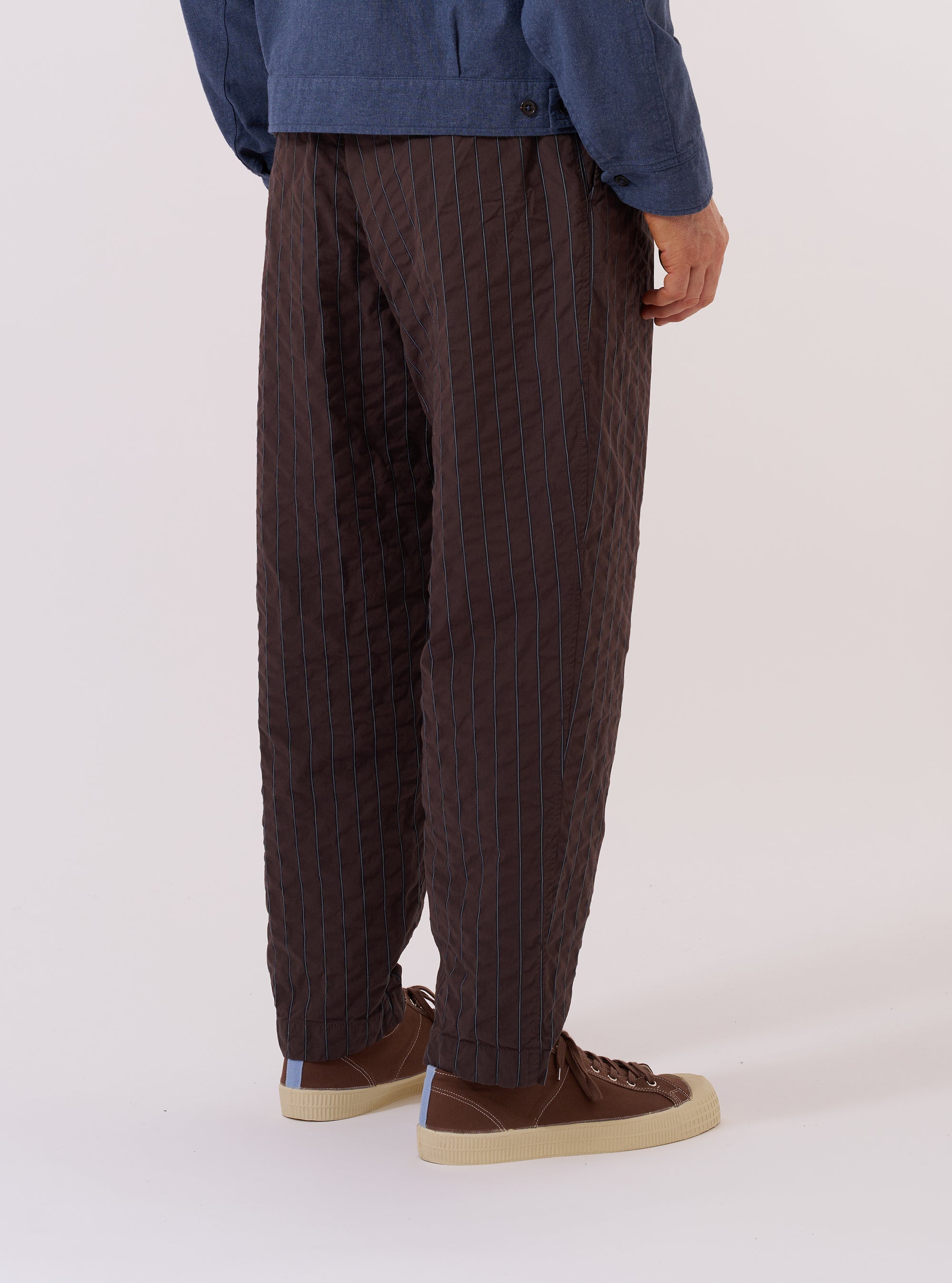 A model wears the Brown Chester Stripe Pleated Track Pant, shown from the waist down from a rear angle against a plain white background. The trousers feature a vertical brown and tan stripe pattern, a relaxed tapered fit, and a single rear patch pocket. The elasticated waistband and the textured cotton fabric are clearly visible as the model stands with one leg slightly forward.
