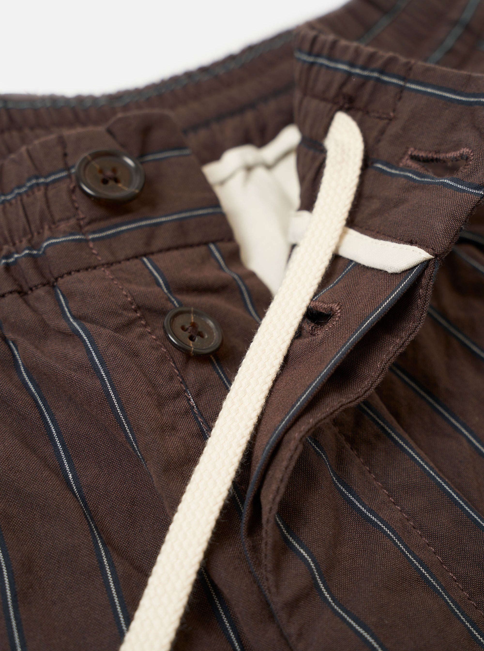 The Brown Chester Stripe Pleated Track Pant is shown from the back as a flat lay against a plain white background. The image highlights the two rear pockets, one with a buttoned flap and the other with a button jetted closure, as well as the elasticated waistband. The brown cotton fabric features a subtle vertical pinstripe pattern throughout.