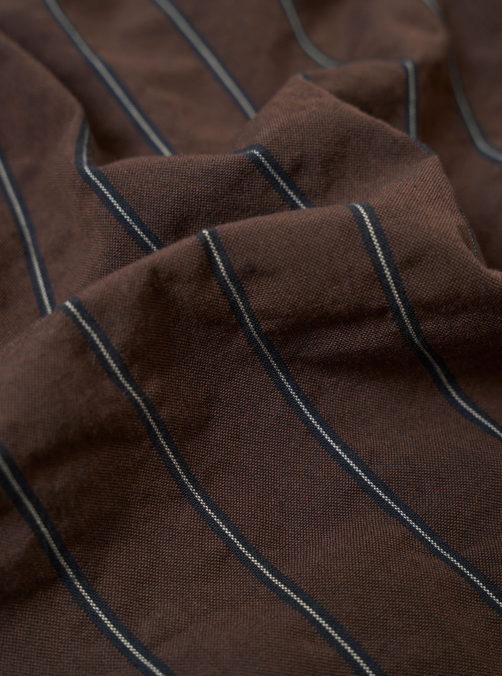 A close-up fabric swatch of the Brown Chester Stripe Pleated Track Pant highlights the texture and pattern of the cotton seersucker material. The image shows a brown base with a refined white pinstripe pattern woven into the puckered fabric.