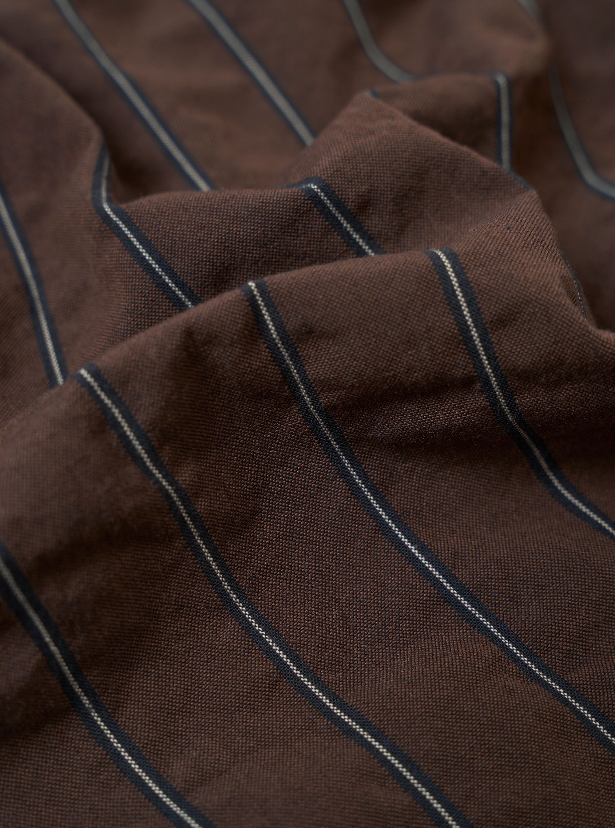 A close-up fabric swatch of the Brown Chester Stripe Pleated Track Pant highlights the texture and pattern of the cotton seersucker material. The image shows a brown base with a refined white pinstripe pattern woven into the puckered fabric.