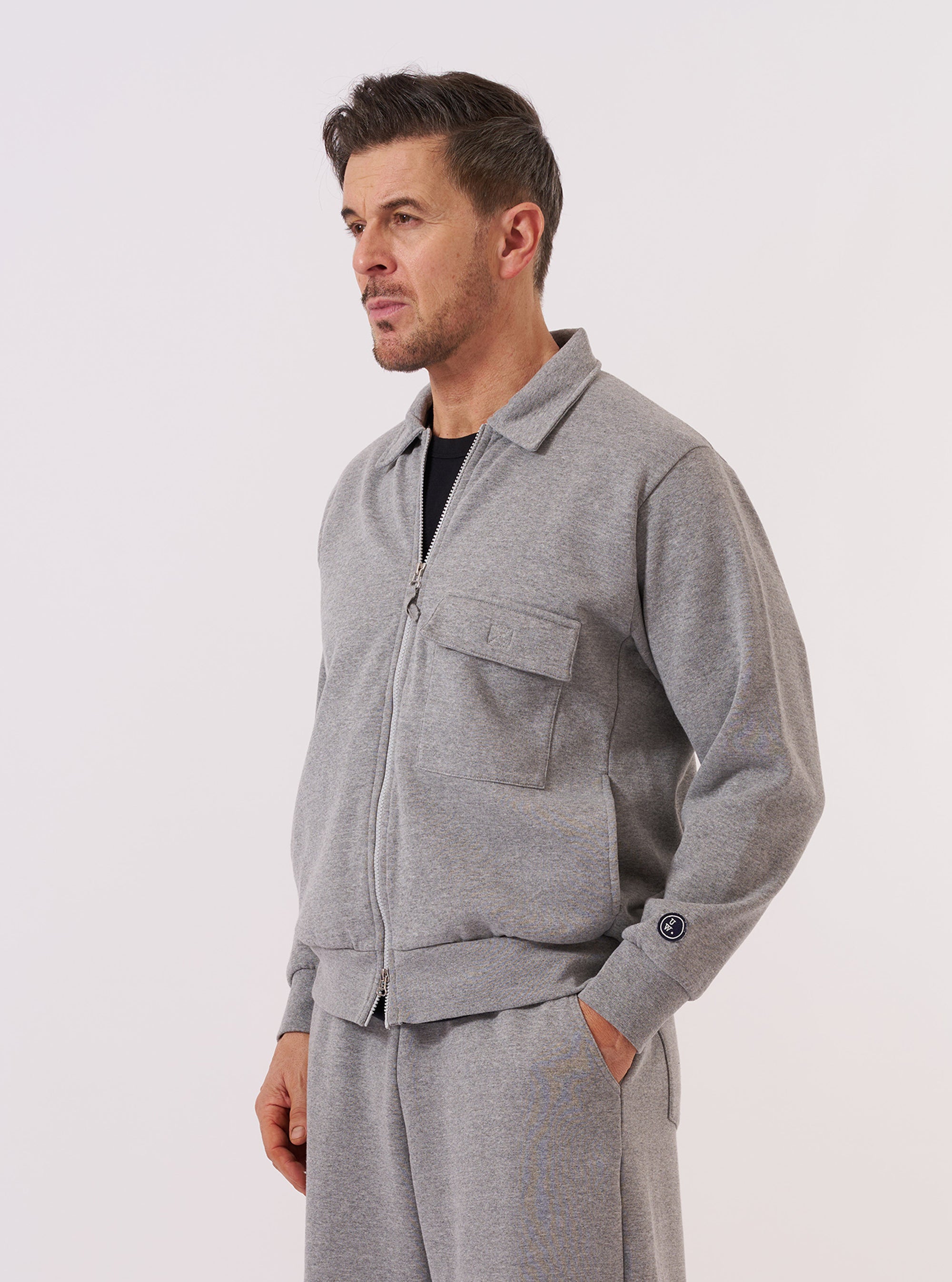 A model wears the Grey Marl Recycled Cotton Blend Jersey Tyler Jacket over a light-colored t-shirt, standing against a plain white background. The jacket features a full-length front zipper, a classic spread collar, a single patch pocket on the chest, and two larger patch pockets at the waist. The grey marl fabric has a soft, heathered appearance.