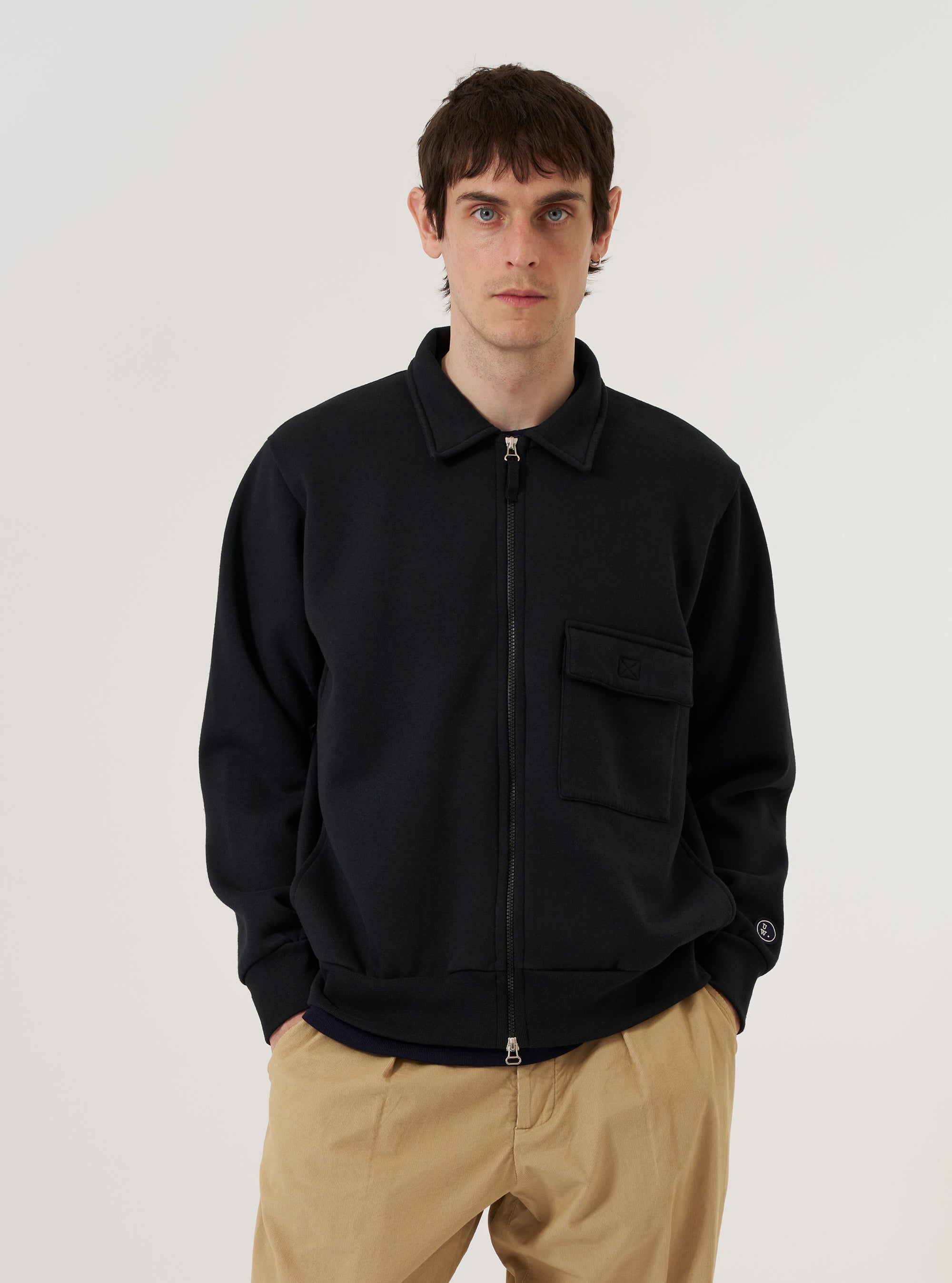 A model wears the Black Recycled Cotton Blend Jersey Tyler Jacket against a plain white background. The jacket features a central two-way zip closure, a self-fabric collar, and a single patch pocket with a velcro fastening on the chest. The black jersey fabric is shown with a relaxed fit, highlighting the ribbed cuffs and hem.