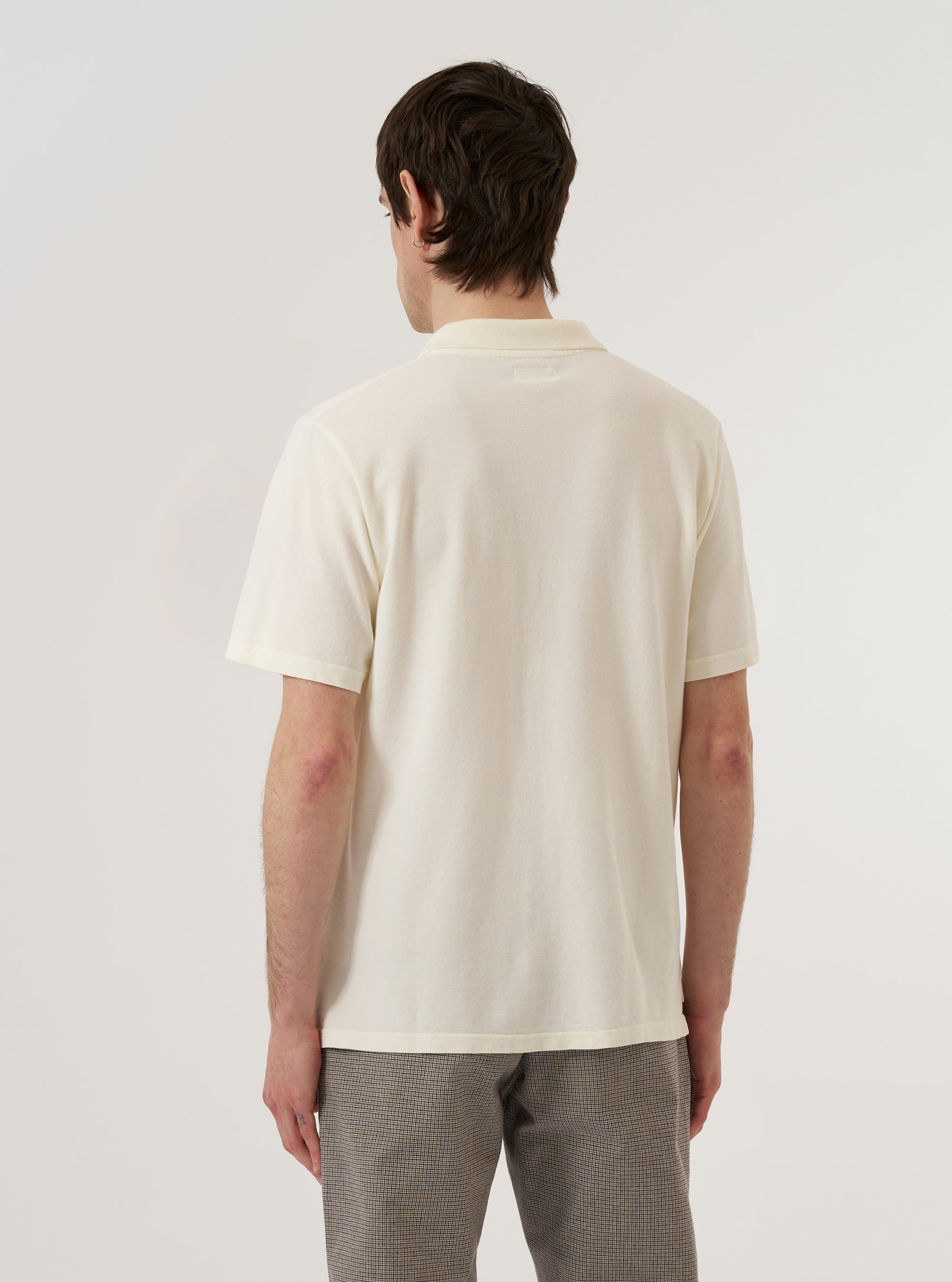 A model wears the Ecru Piquet Vacation Polo, shown from a rear three-quarter angle against a plain white background. The polo features a relaxed fit, short sleeves, and a one-piece collar. The textured ecru piquet cotton fabric and a straight hem are clearly visible as the model stands facing away from the camera.