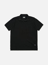 The Black Piquet Vacation Polo is shown as a flat lay against a plain white background. The polo features a single-piece open collar, a short-button placket, and a single patch pocket on the left chest. The black cotton fabric has a visible textured piquet weave.