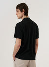 A model is wearing the Black Piquet Vacation Polo against a plain white background. The polo features a textured piquet knit fabric, short sleeves, and a relaxed vacation-style collar. The model is shown from the waist up, highlighting the fit and the weave of the black fabric.