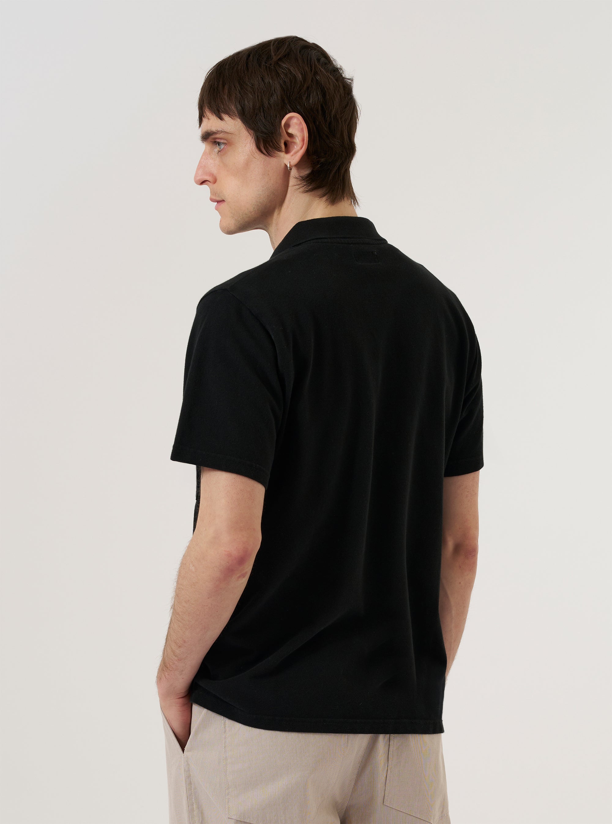 A model is wearing the Black Piquet Vacation Polo against a plain white background. The polo features a textured piquet knit fabric, short sleeves, and a relaxed vacation-style collar. The model is shown from the waist up, highlighting the fit and the weave of the black fabric.