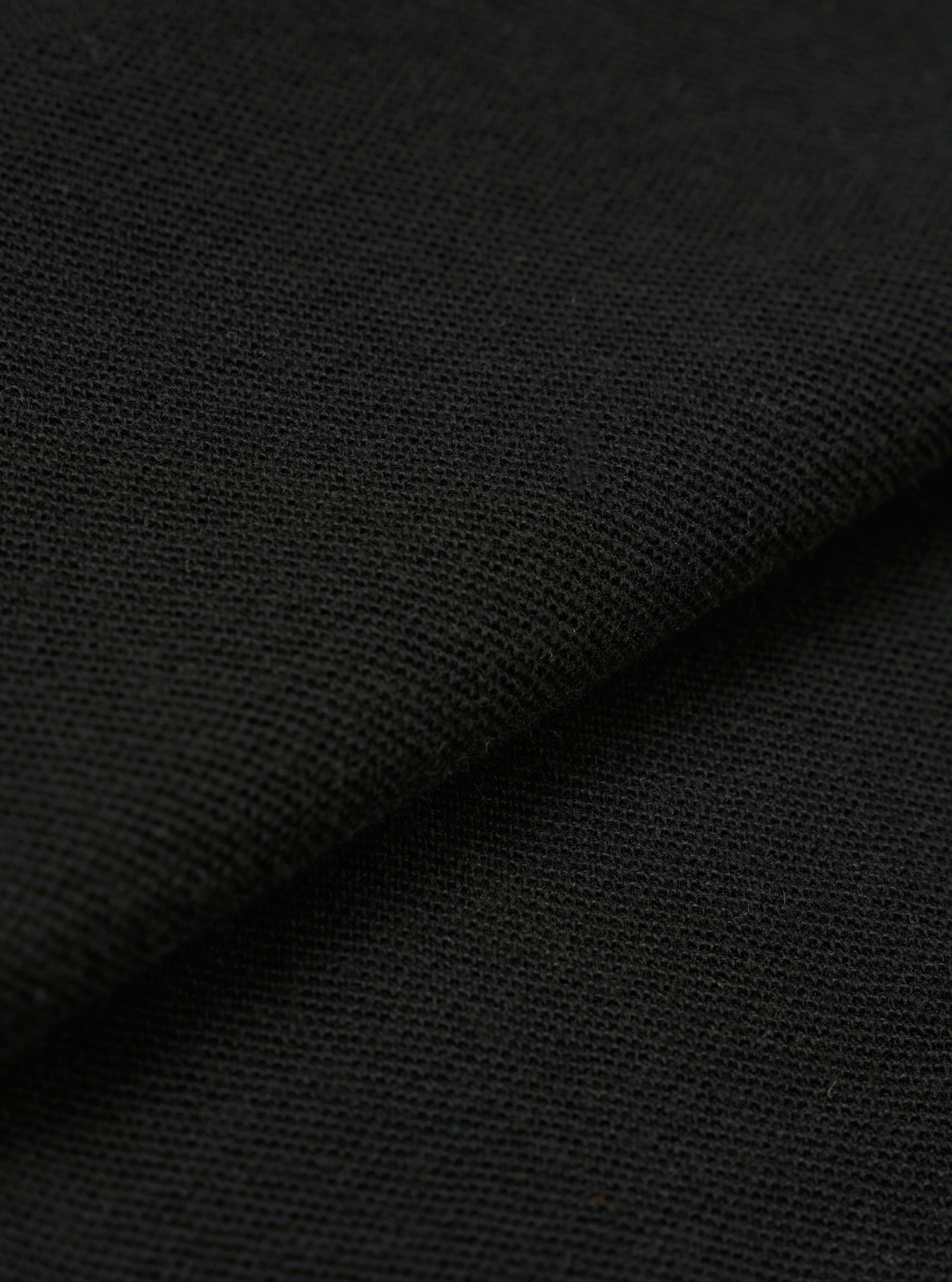 A close-up fabric swatch of the Black Piquet Vacation Polo highlights the textured, waffle-like weave of the black piquet cotton material. The image provides a detailed view of the fabric's tactile surface and solid black color.