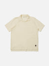 The Ecru Piquet Vacation Polo is shown as a flat lay against a white background. The polo features a one-piece open collar, a single chest patch pocket on the left side, and short sleeves. The fabric has a distinct textured piqué weave in a solid ecru color.