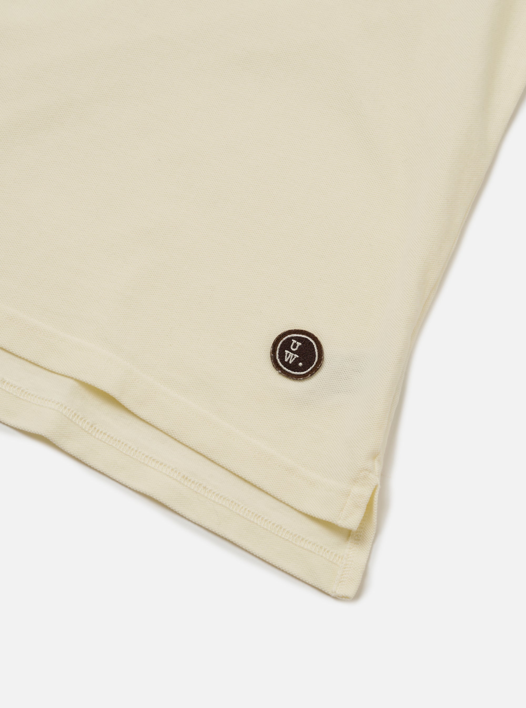 The Ecru Piquet Vacation Polo is shown in a detail view as a flat lay against a plain white background. The image focuses on the lower portion of the garment, highlighting the textured ecru piqué fabric, the side vents, and the stepped hem. A small brand logo patch is visible near the bottom hem.