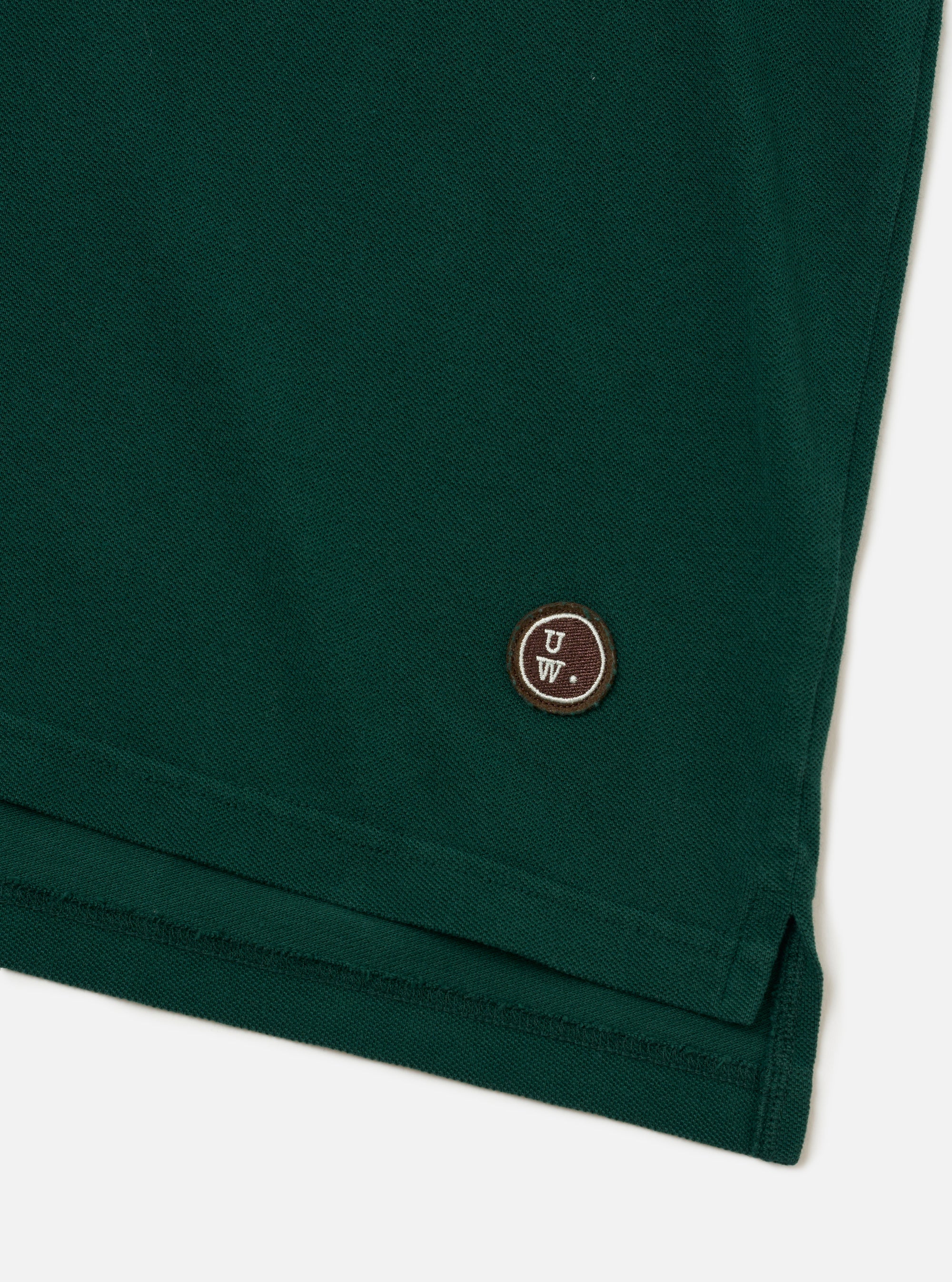 The Green Piquet Vacation Polo is shown in a detail view as a flat lay against a white background. The image focuses on the lower portion of the garment, highlighting the clean hemline and the textured green piquet cotton fabric.