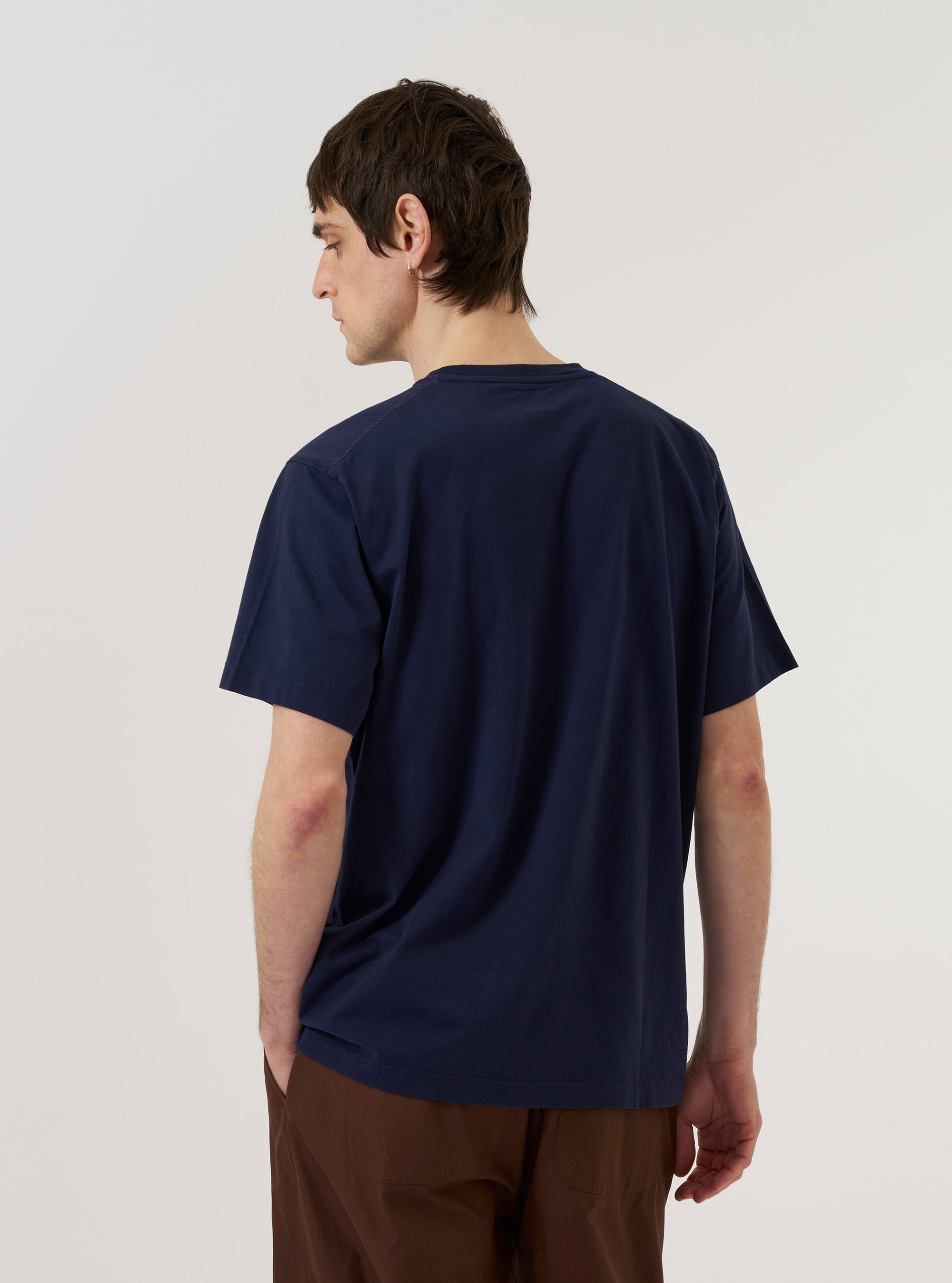 A model wears the Bright Navy Organic Jersey Print Pocket Tee, shown from the back against a plain white background. The t-shirt features a regular fit and a military-inspired logo print on the back. The garment is made from a bright navy organic cotton jersey.