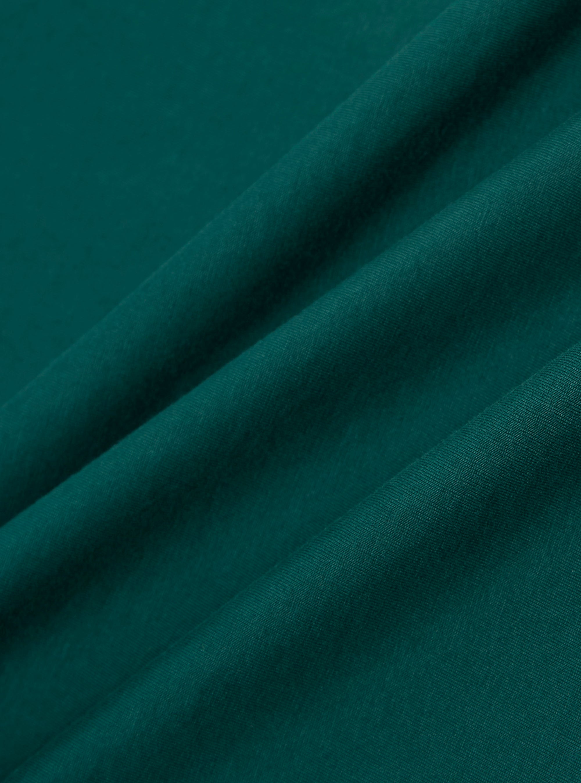 A close-up fabric swatch of the Olive Organic Jersey Print Pocket Tee displays the olive green color and soft, fine-knit texture of the organic cotton jersey material. The fabric is shown in detail, highlighting its consistent matte finish and weave.
