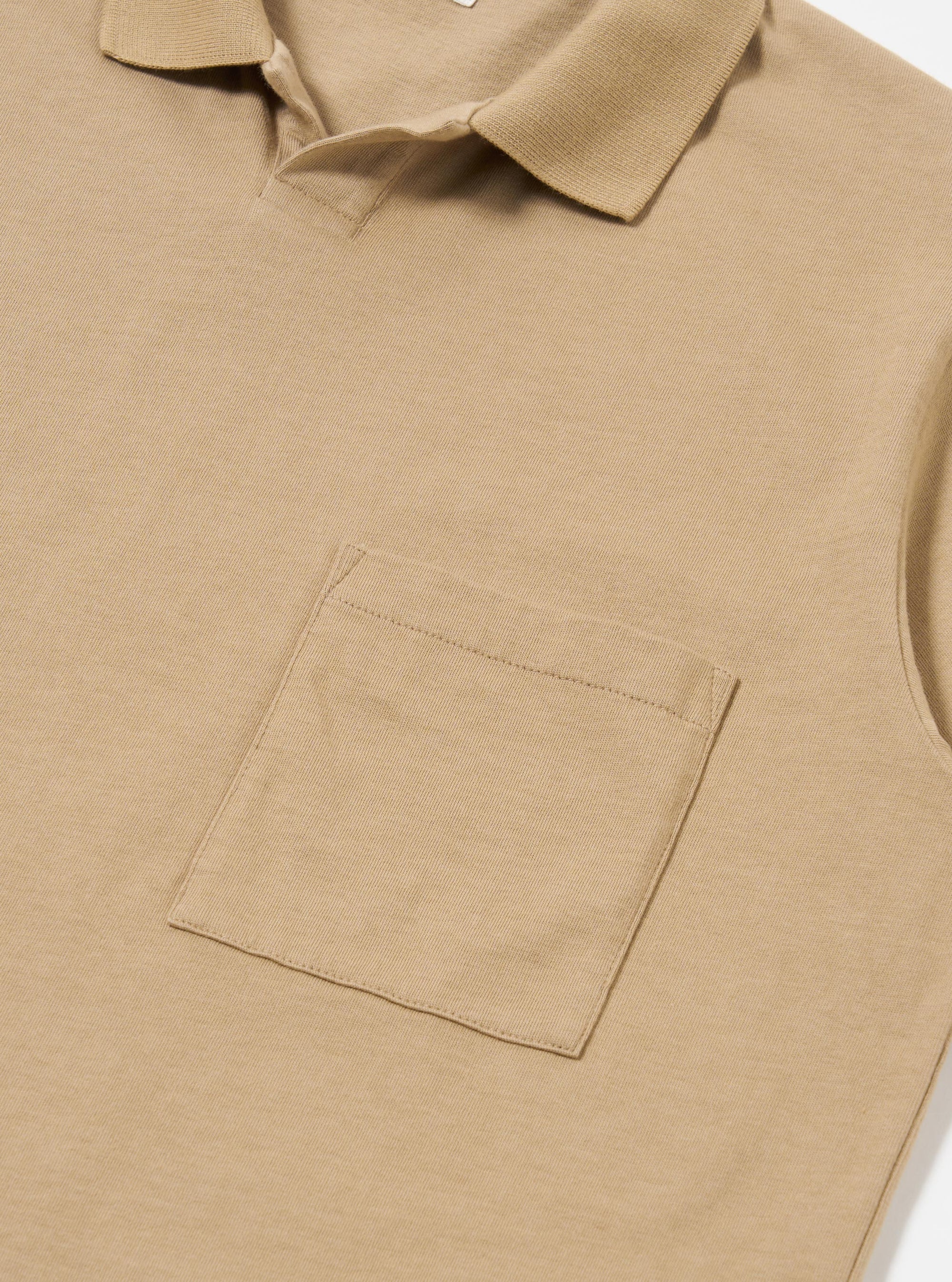 The Sand Rugby Jersey Oversized Vacation Polo is shown from the back as a flat lay against a white background. The image highlights the stepped hem, where the back of the polo is longer than the front, and the small side vents. The sand-colored organic cotton fabric has a smooth, jersey texture.