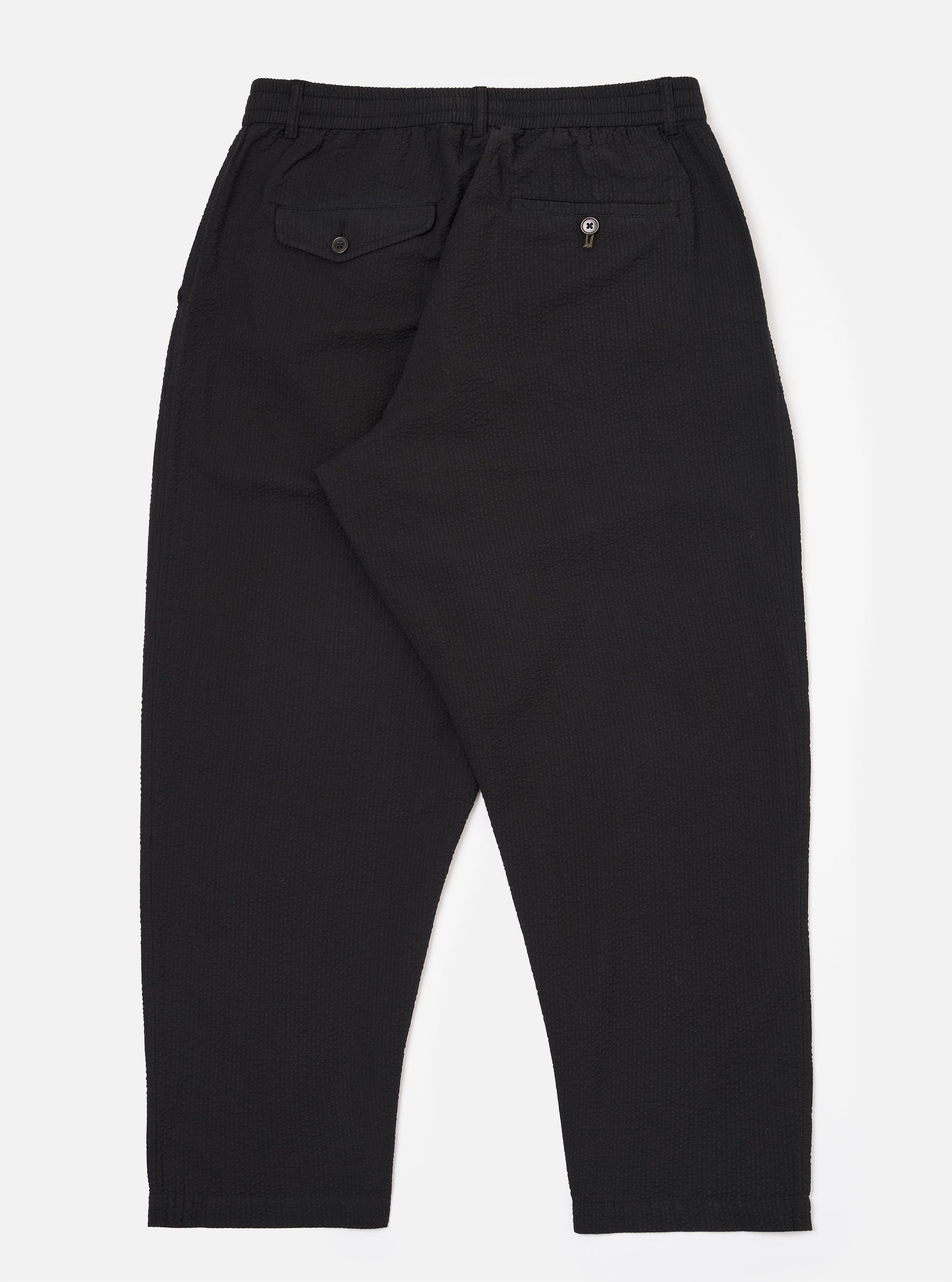 The Black Merida Seersucker Pleated Track Pant is shown from the back as a flat lay against a plain white background. The image highlights the elasticated waistband and two rear pockets, including one with a buttoned flap and one buttoned jetted pocket. The black cotton seersucker fabric has a visible puckered texture.