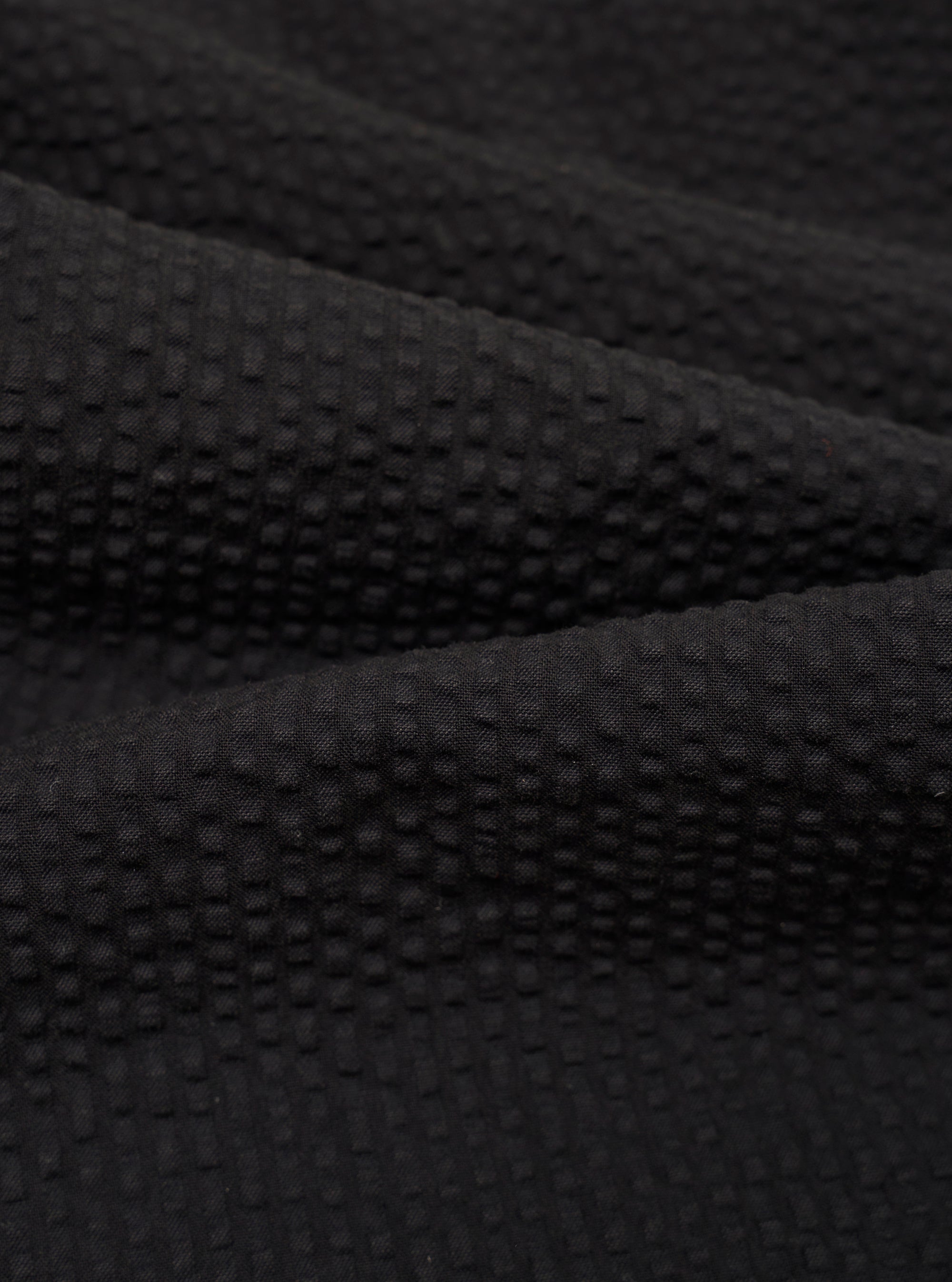 A close-up swatch image of the Black Merida Seersucker Military Chino fabric highlights the garment's black color and the distinct puckered texture of the cotton seersucker material. The image focuses entirely on the textile detail, providing a clear view of the fabric's weave.