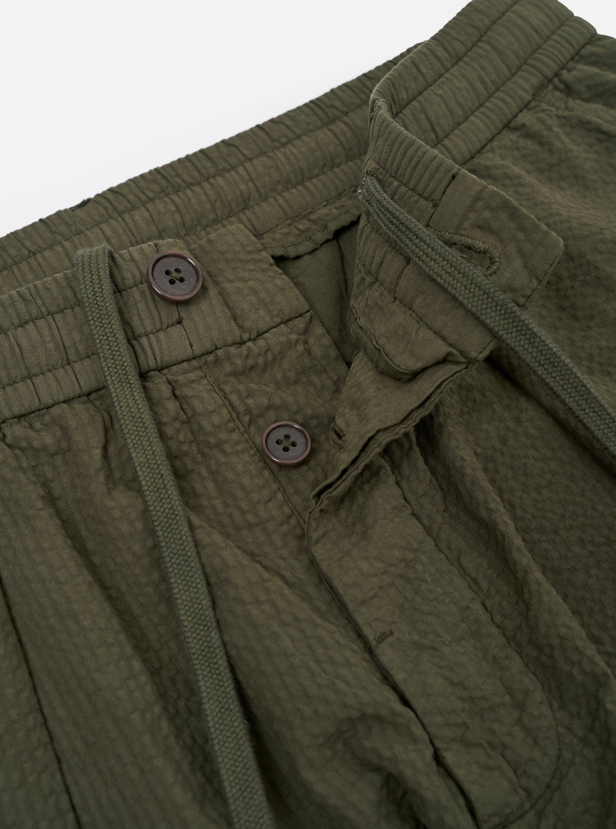 The Olive Merida Seersucker Pleated Track Pant is shown from the back as a flat lay against a plain white background. The image highlights the elasticated waistband and the two rear pockets, including one buttoned flap pocket on the left and one button jetted pocket on the right. The olive green fabric features a distinct puckered seersucker texture.