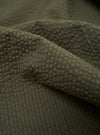 A close-up fabric swatch of the Olive Merida Seersucker Pleated Track Pant highlights the olive green color and the distinct puckered texture of the cotton seersucker material.