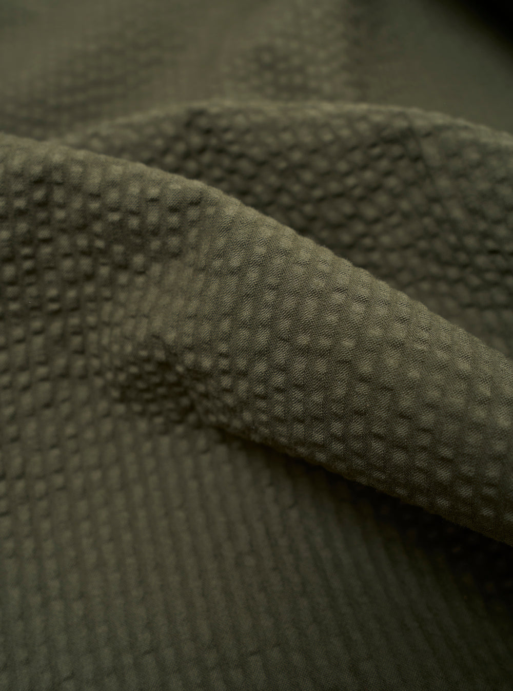 A close-up fabric swatch of the Olive Merida Seersucker Pleated Track Pant highlights the olive green color and the distinct puckered texture of the cotton seersucker material.