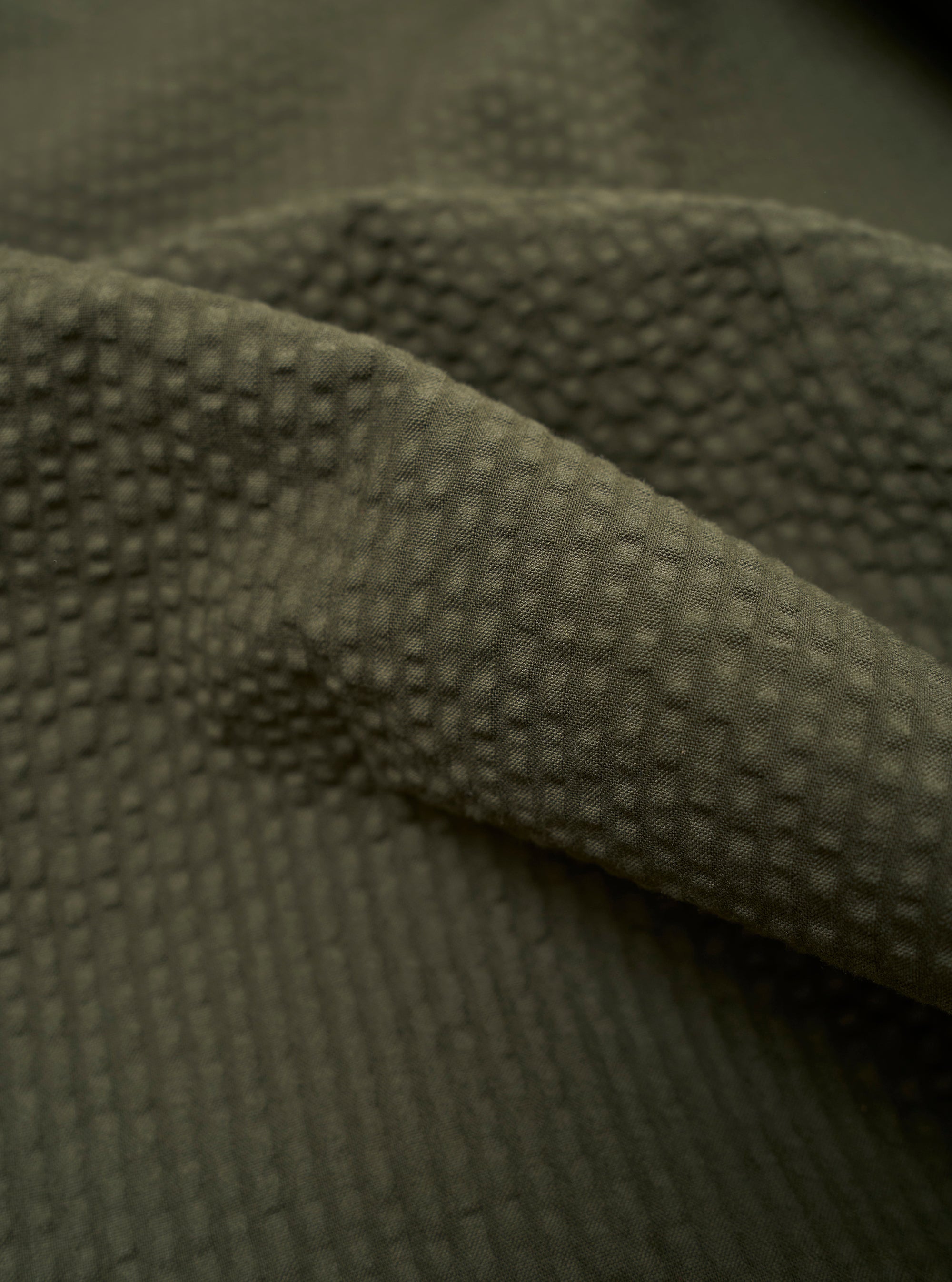 A close-up fabric swatch of the Olive Merida Seersucker Pleated Track Pant highlights the olive green color and the distinct puckered texture of the cotton seersucker material.