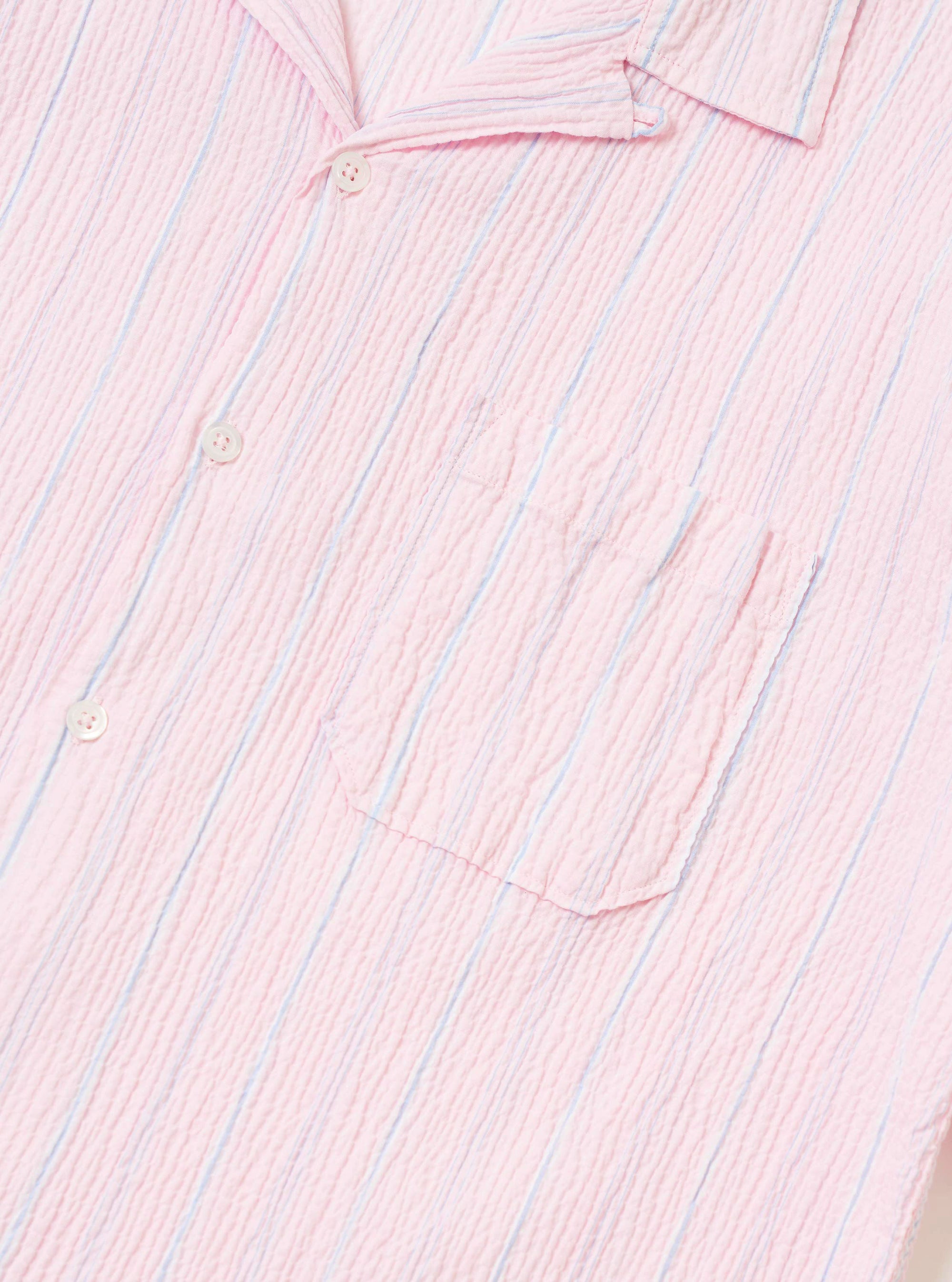 The Pink Lily Stripe Road Shirt is shown from the back, worn by a model. The shirt features a camp collar and short sleeves, with a vertical stripe pattern in shades of pink, white, and red. The model is also wearing olive green trousers. The background is a plain, neutral light gray.