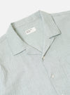 The Green Seersucker Road Shirt is worn by a model against a plain, neutral background. The shirt features a relaxed camp collar, a button-front closure, and a single chest pocket. The short-sleeve garment is made from a green cotton fabric with a distinct, puckered seersucker texture. The model is shown from the front, wearing the shirt untucked.