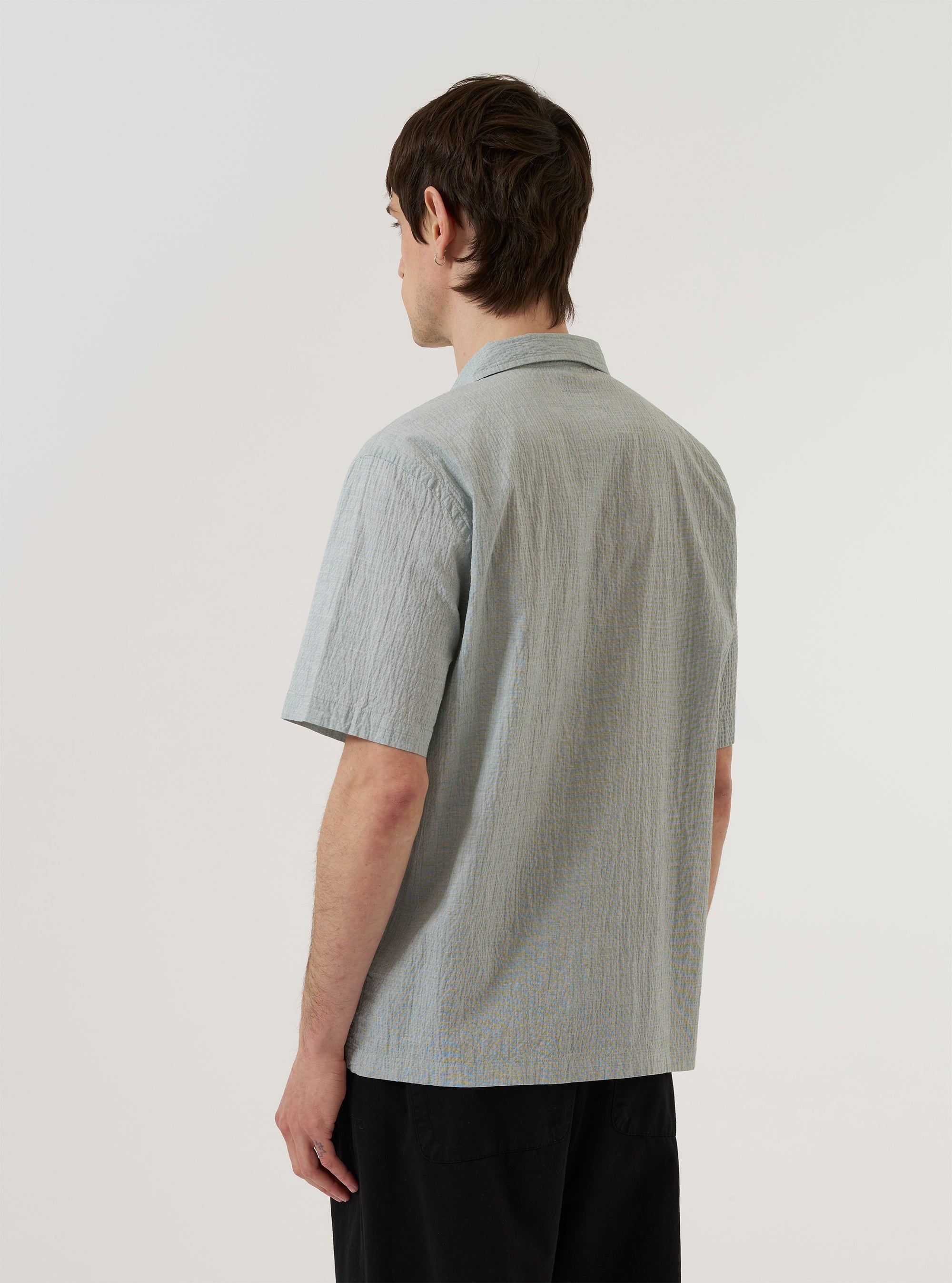 A model wears the Green Seersucker Road Shirt against a plain white background, shown from the chest down. The short-sleeve shirt is worn open over a white t-shirt, highlighting the textured seersucker fabric and the straight hem. The model's hands are tucked into the pockets of olive green trousers.